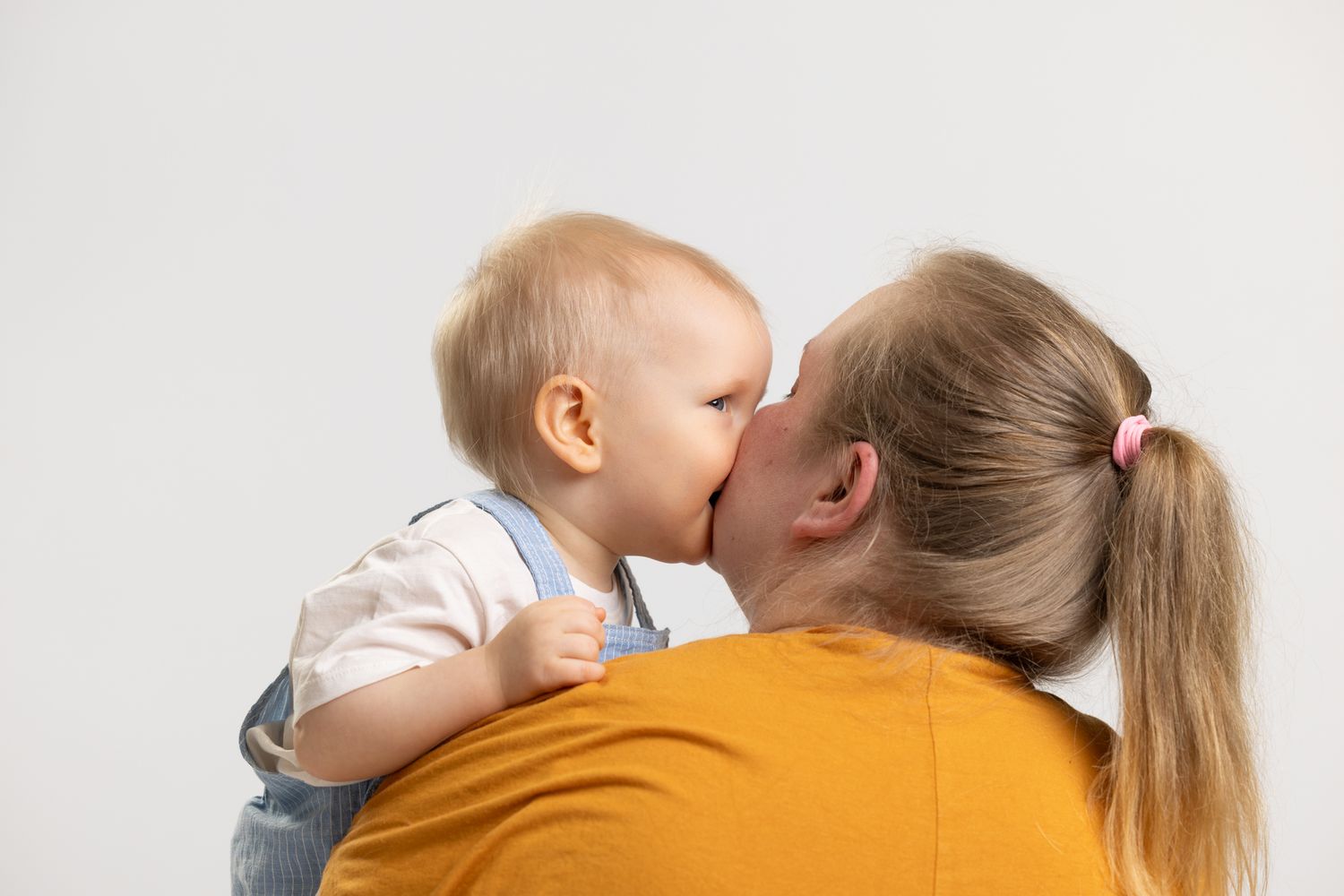 A baby kissing a woman with a blonde ponytail wearing a yellow shirt against a white background.