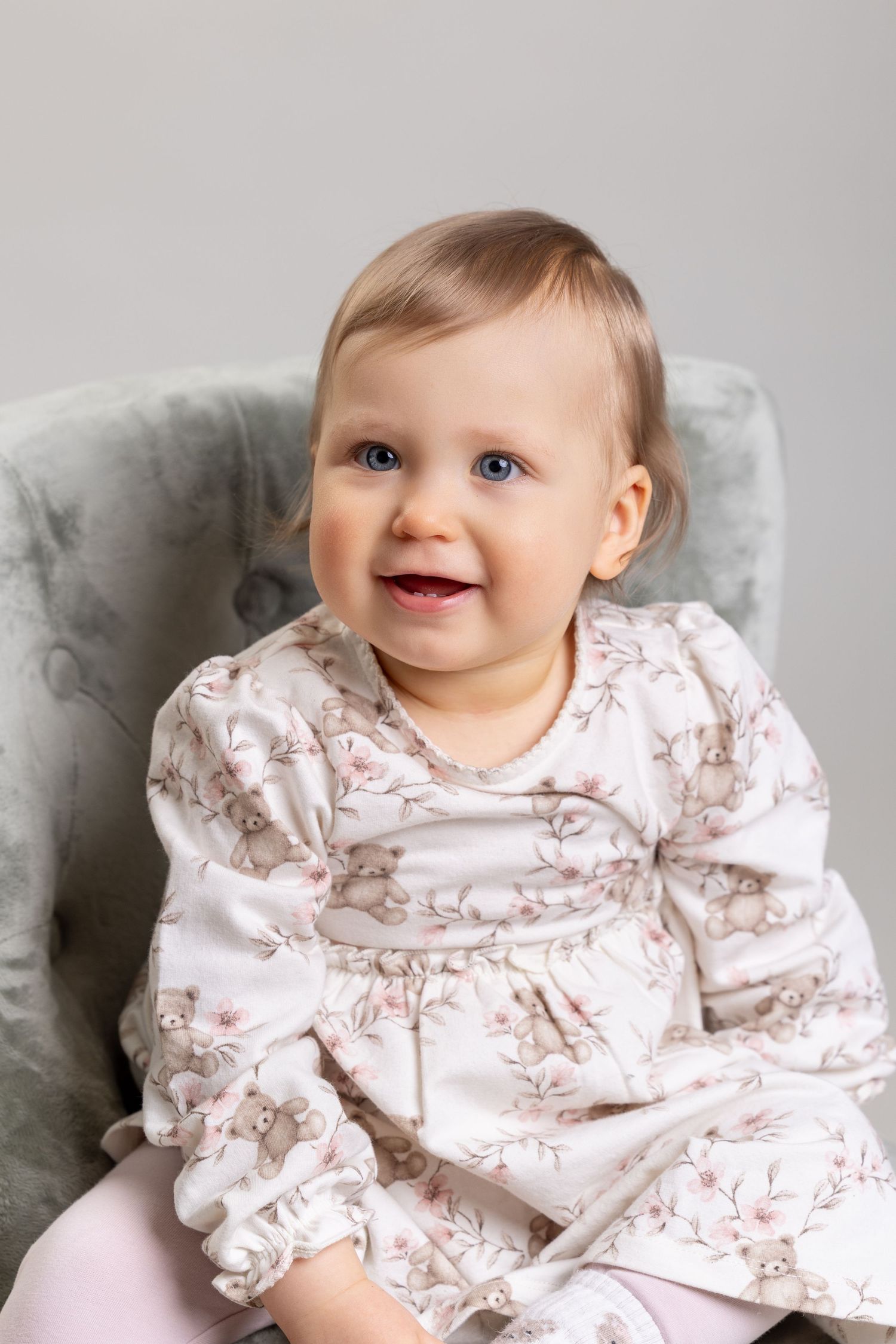 Smiling baby girl in a floral dress sitting in a gray chair, looking happy and cheerful.