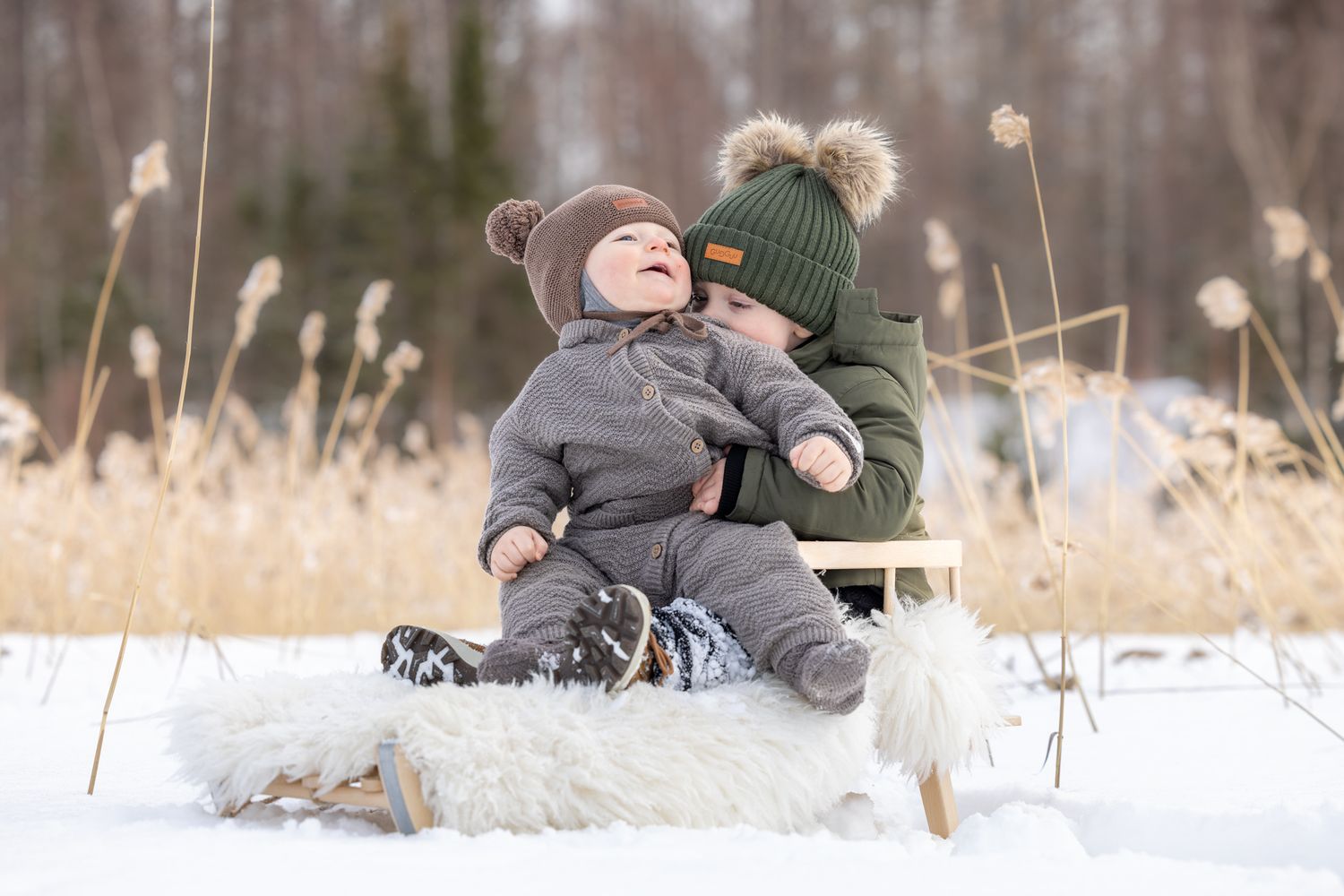 Two young children in winter hats laugh on a wooden sled in snowy landscape.