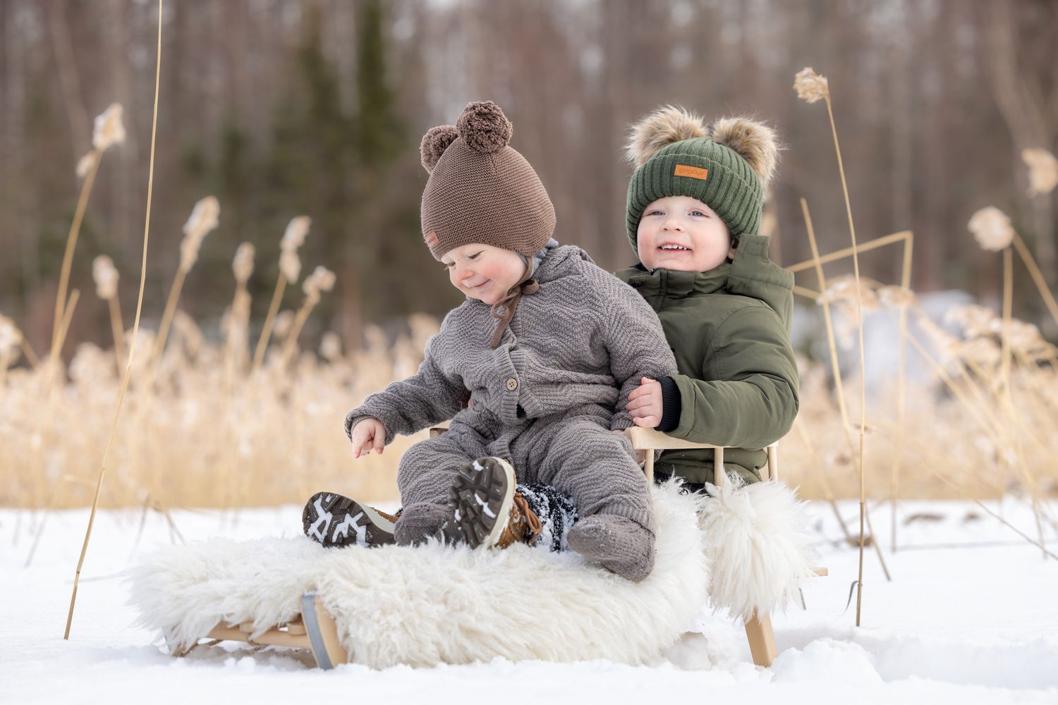 Two toddlers in winter snowsuits sit on a sled in snowy landscape, wearing knit bear-ear hats, smiling outdoors.