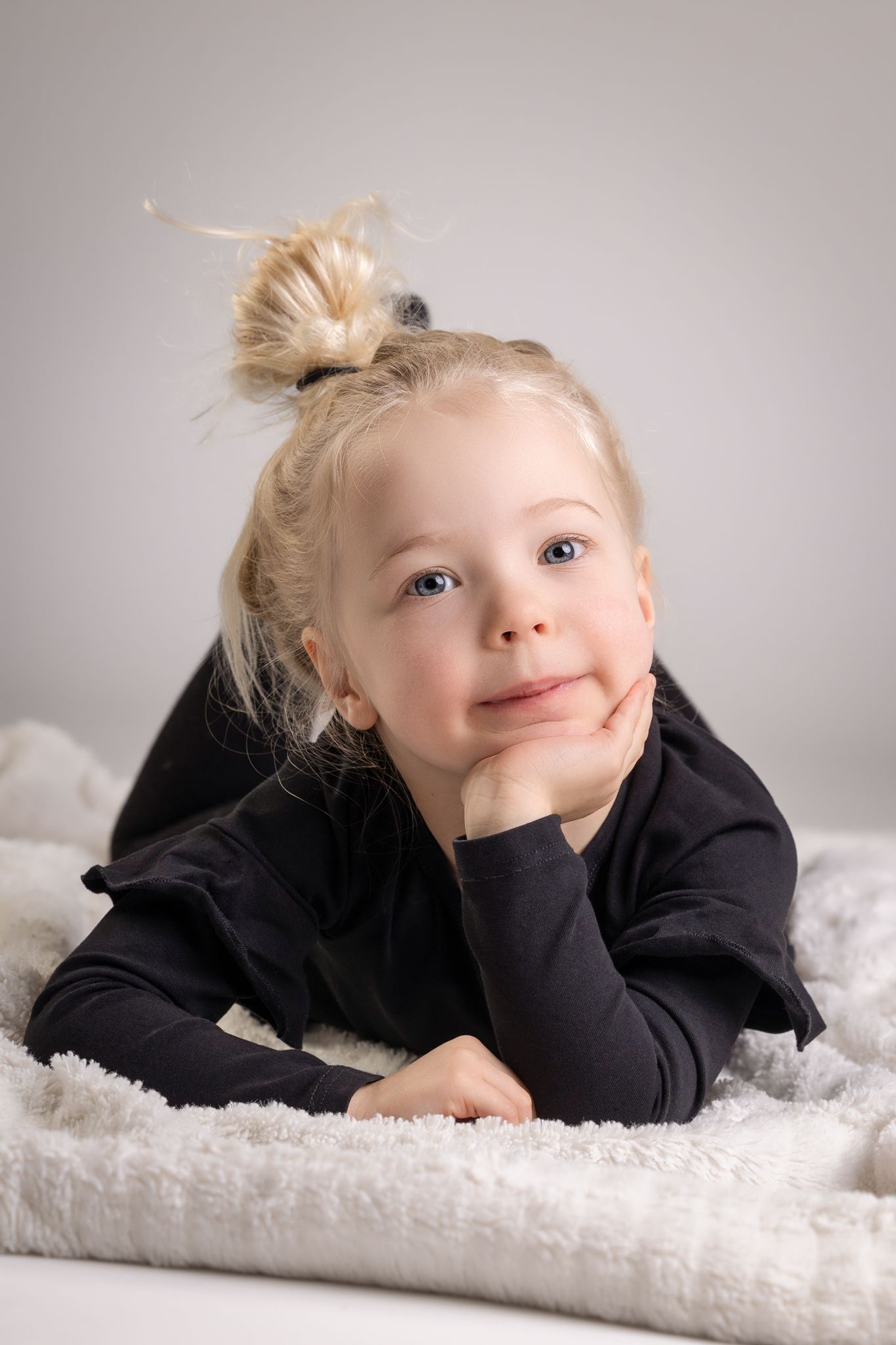 Adorable blonde toddler girl in black outfit lying on fluffy blanket, resting chin on hand with a cute smile.