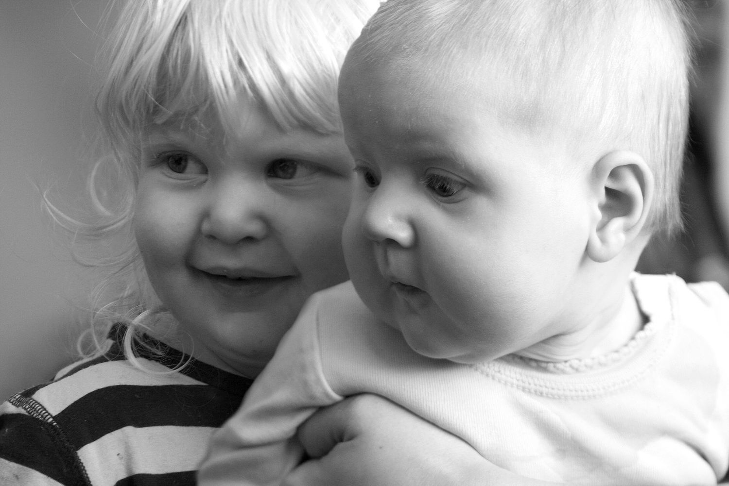 Black and white photo of two young blonde siblings, an older toddler smiling and holding a happy baby.