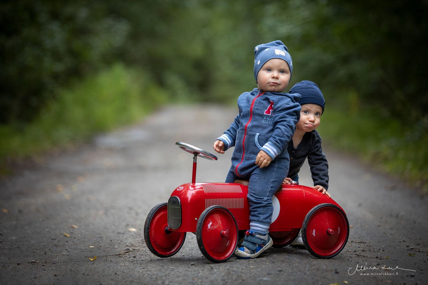 Kaksi pientä poikaa punaisen potkuauton päällä soratiellä.