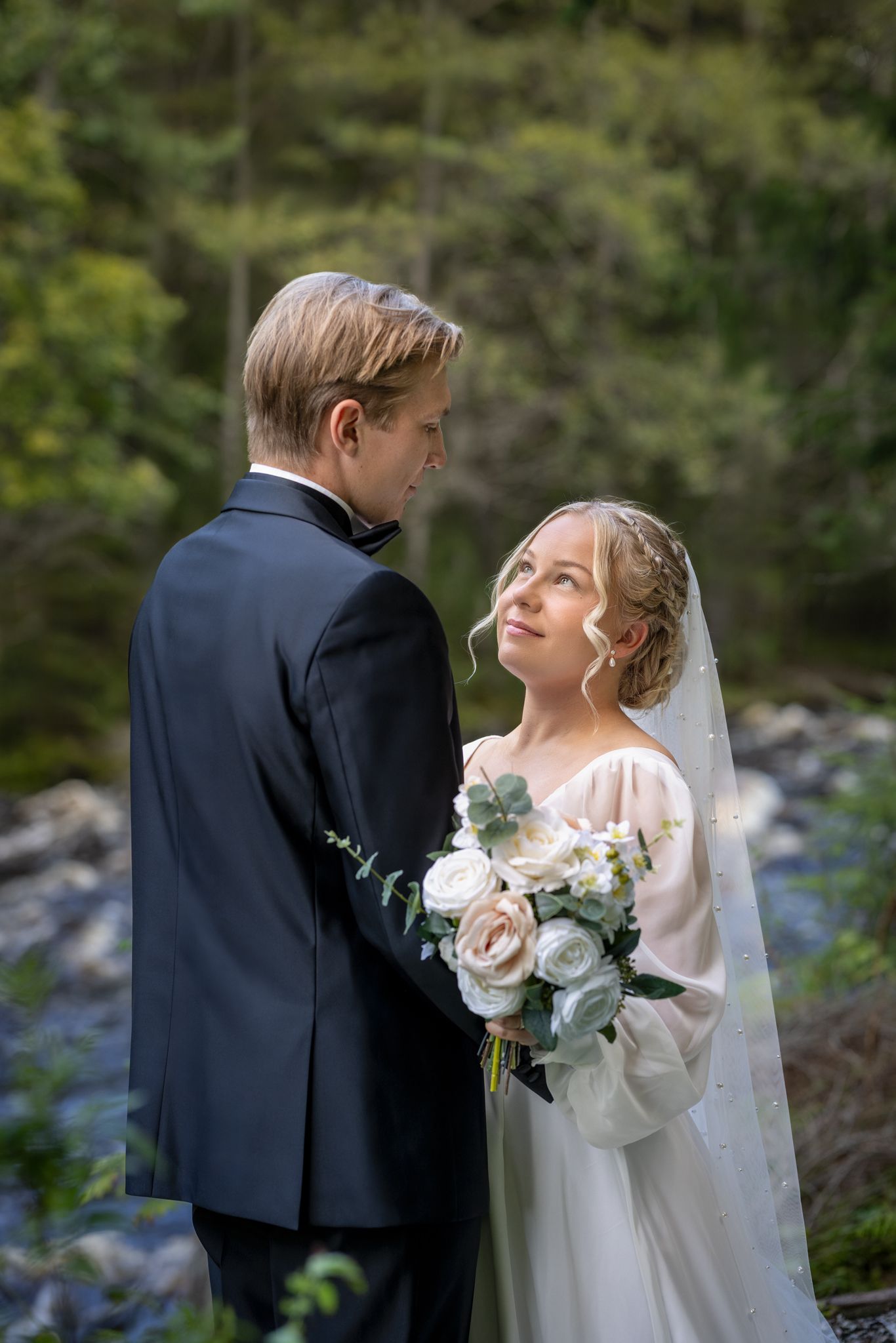 Bride and groom share a tender moment outdoors, she holds white roses bouquet wearing veil, he wears dark suit.