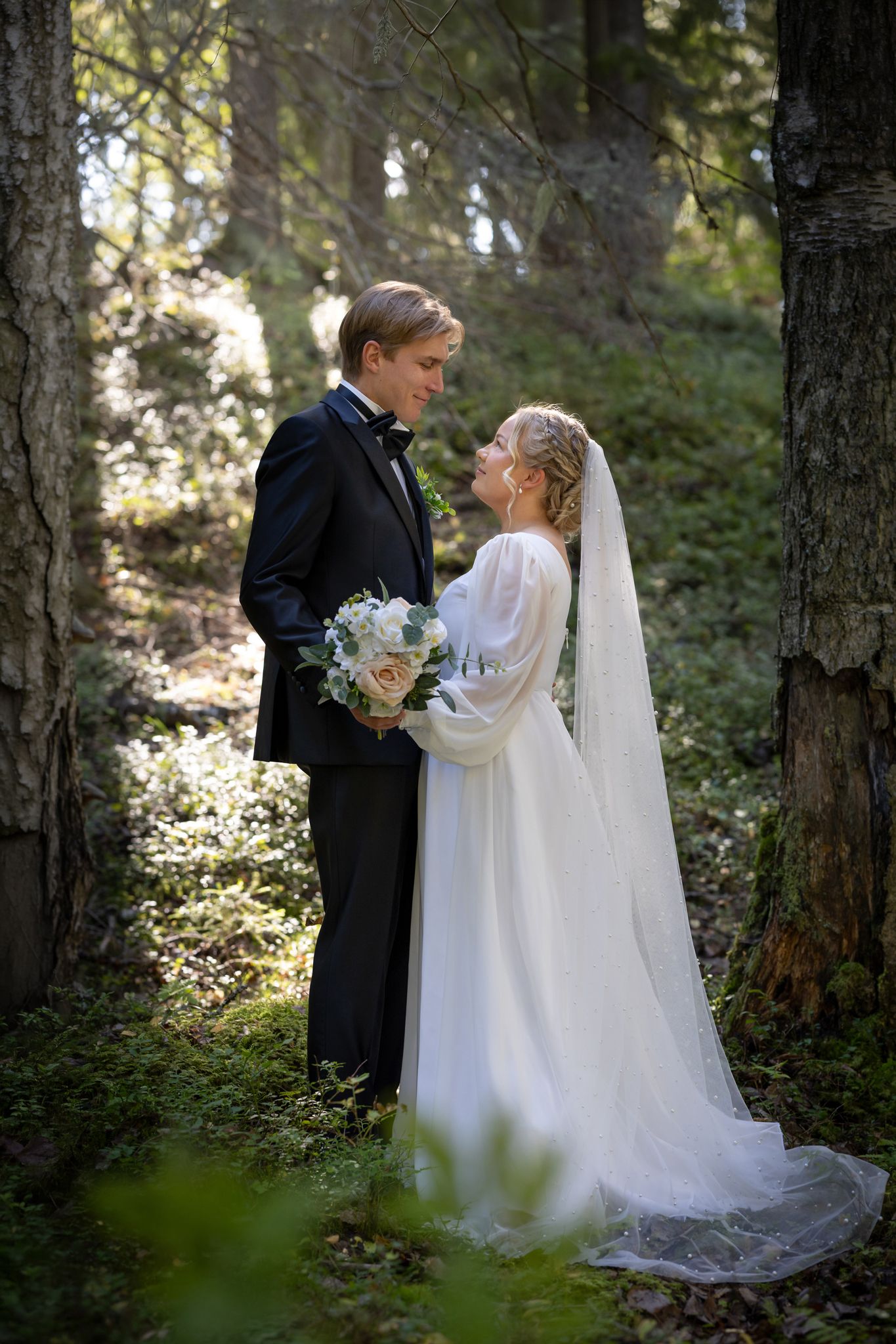 Bride and groom share a romantic moment in a sunlit forest, she in a white gown with veil, he in a black tuxedo.