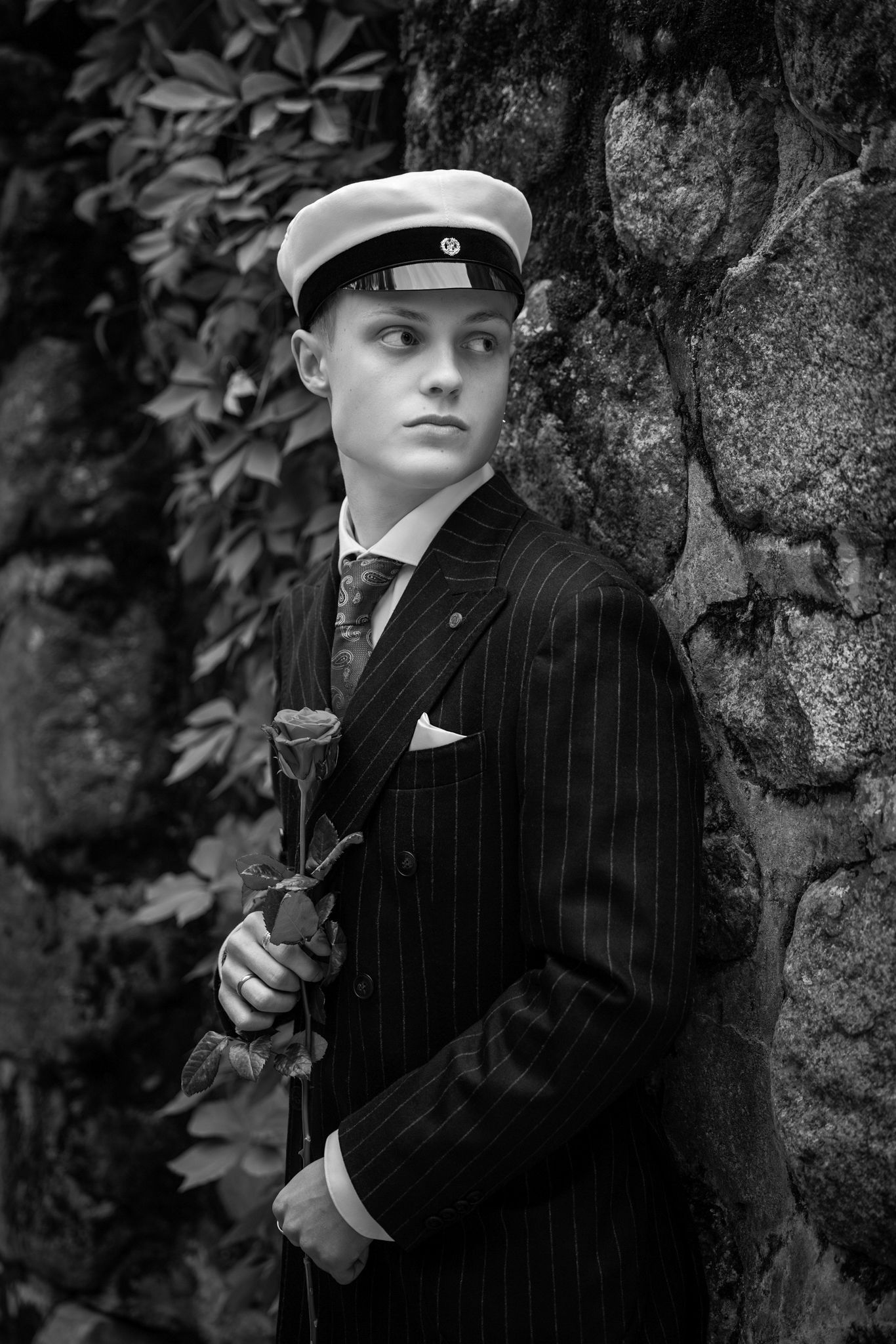 Young man in pinstripe suit and white cap holds a rose against a stone wall in black and white portrait.