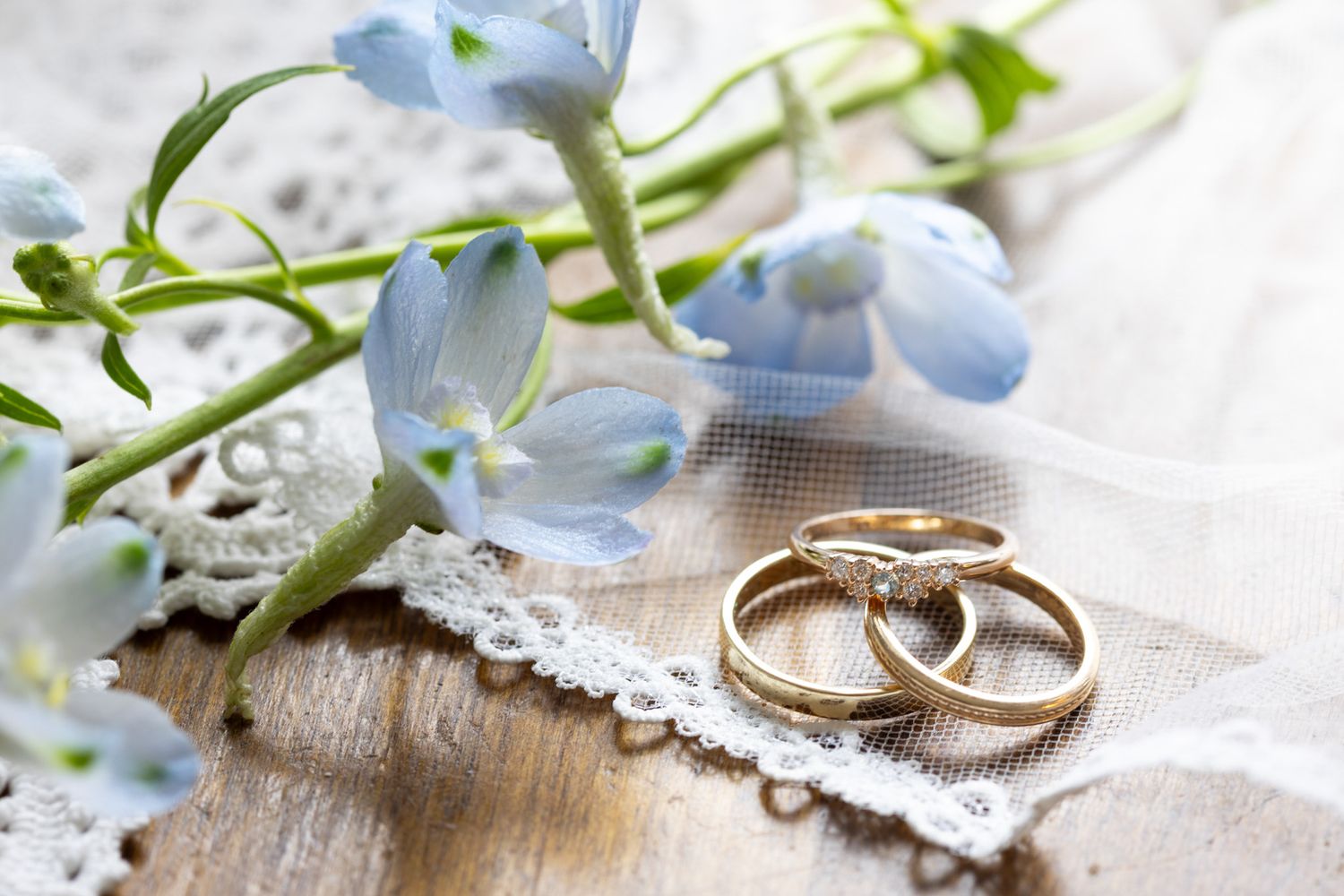 Two gold wedding rings rest on lace beside blue flowers and white veil fabric on a rustic wooden surface.