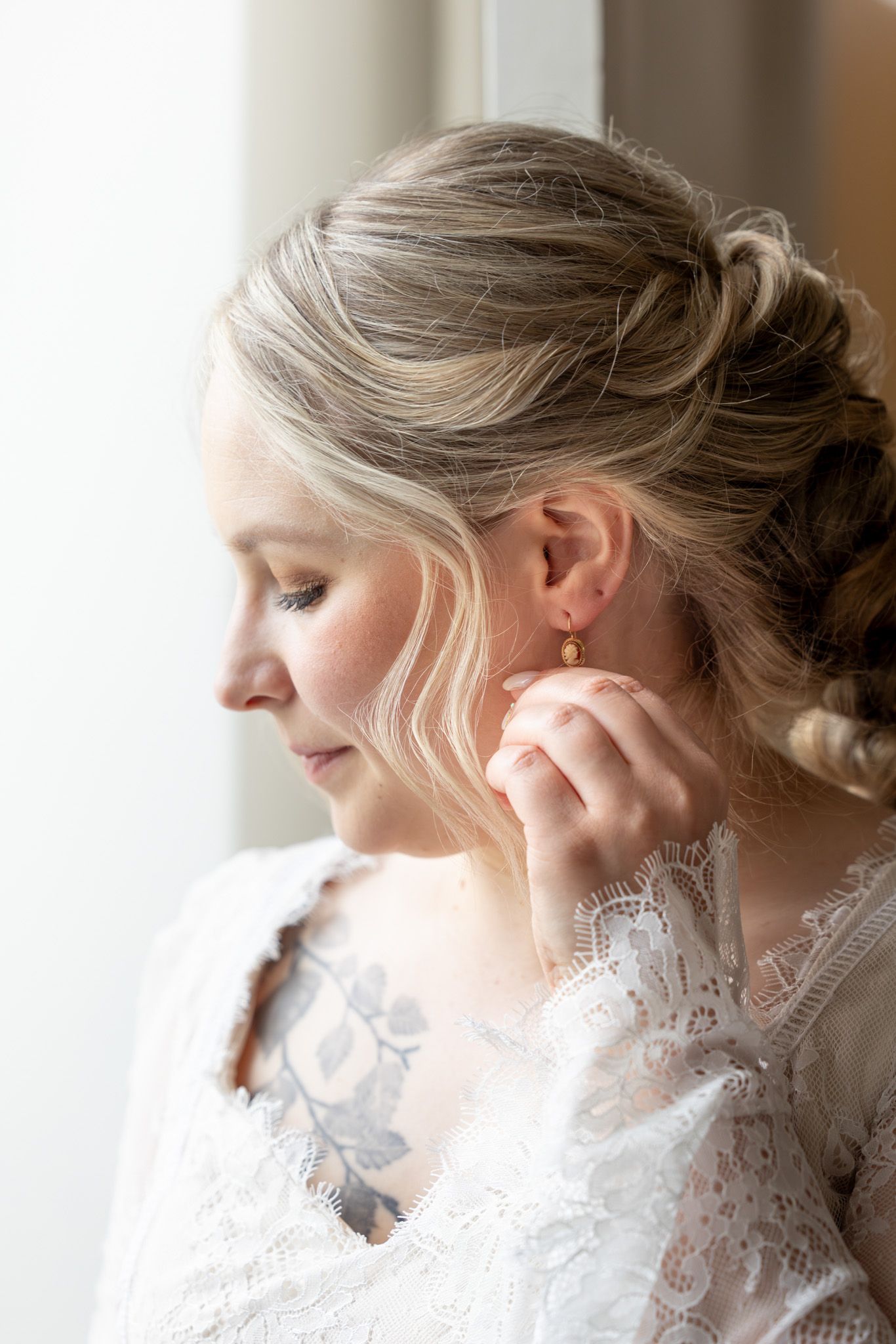 Blonde bride in lace dress adjusting gold earring near window on wedding day.