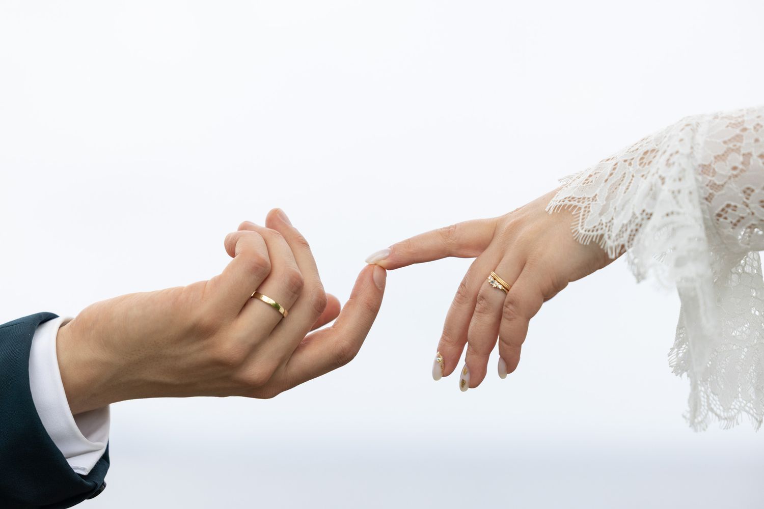 Bride and groom touching fingers, both wearing gold wedding rings, with lace wedding dress sleeve visible.