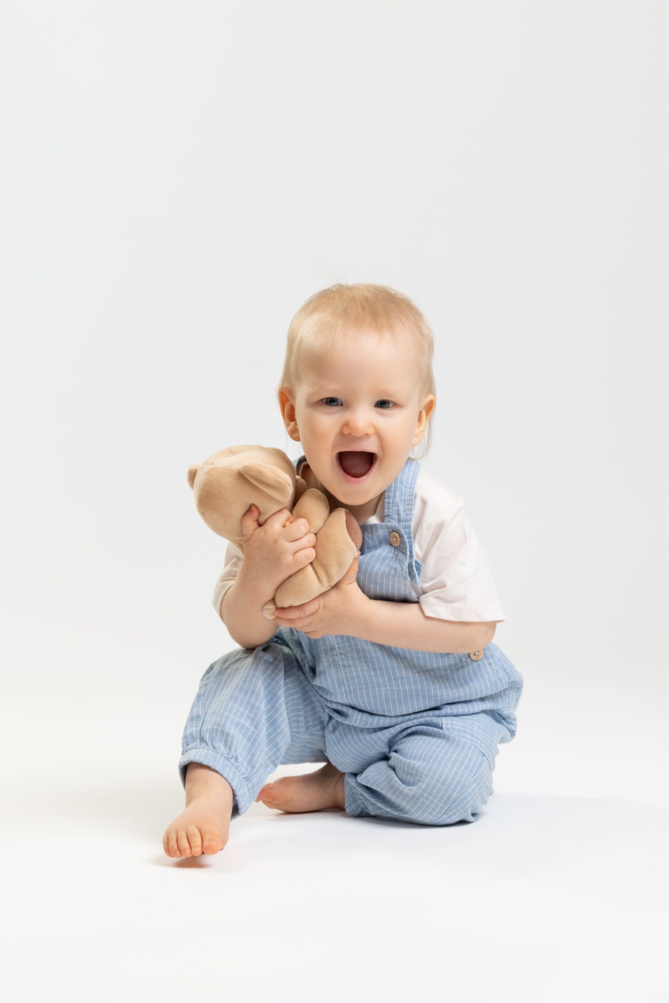 Happy baby in blue overalls hugging a teddy bear, sitting on white background.