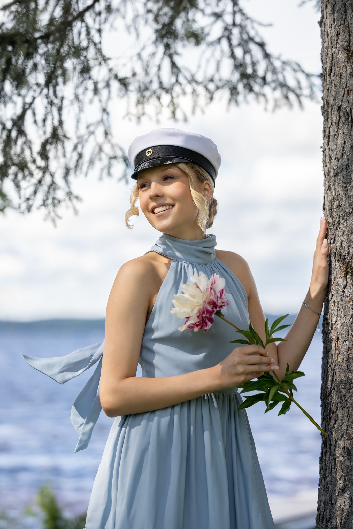 Smiling blonde woman in blue dress and sailor cap holds a peony flower while leaning against a tree by the water.