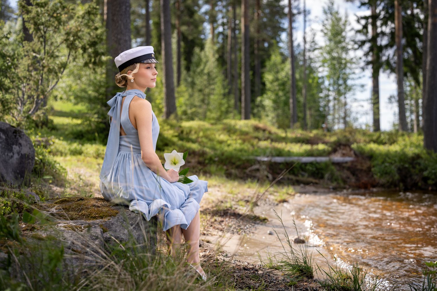 Young girl in blue dress and hat sits on a log by a forest stream holding white flowers.