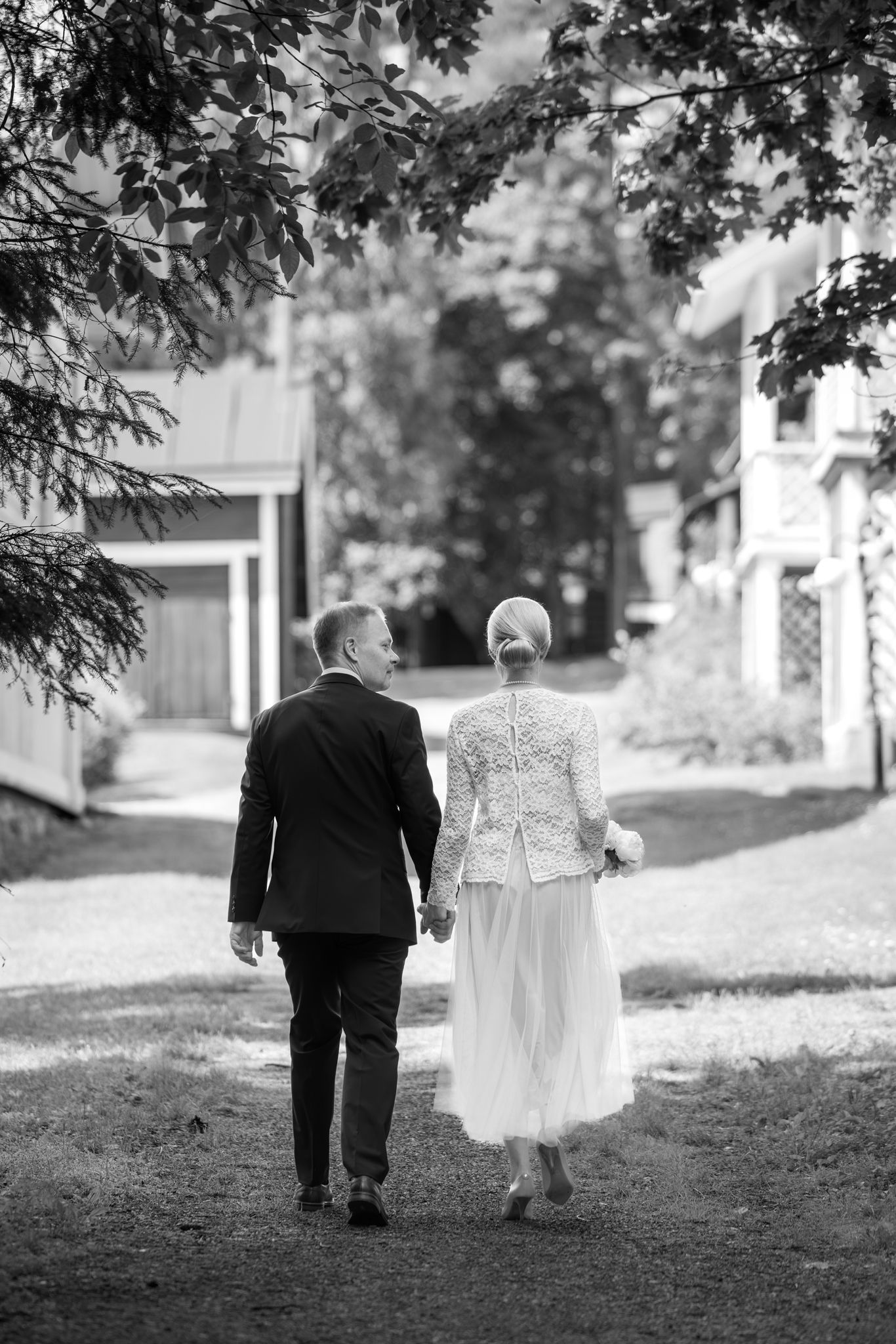 Black and white photo of a bride and groom walking hand in hand down a tree-lined path on their wedding day.