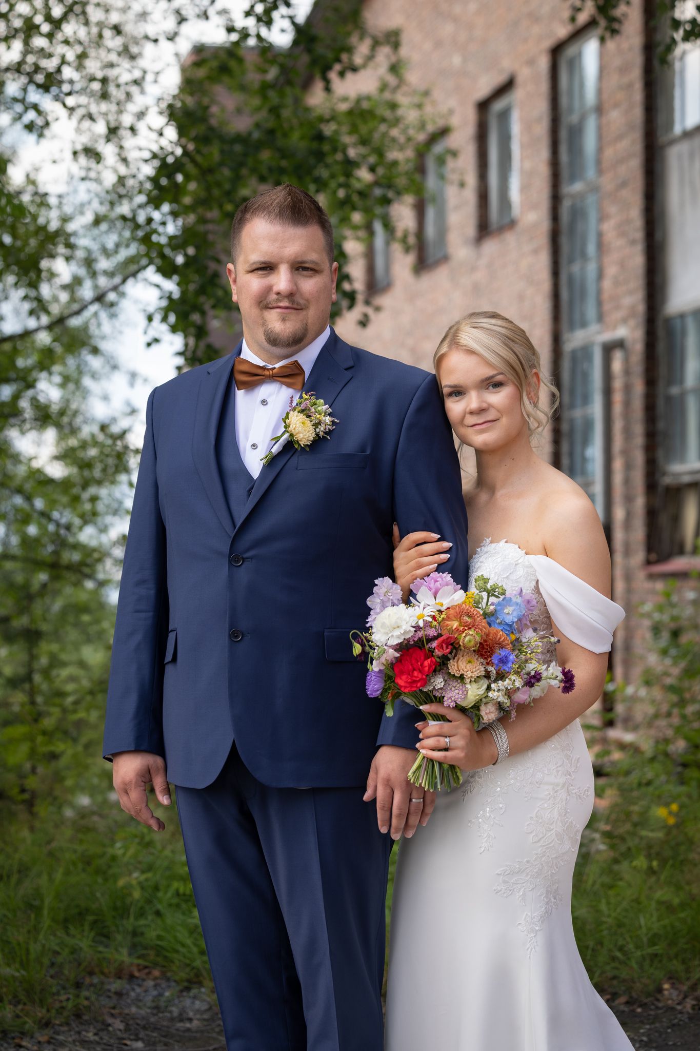 Bride and groom posing outdoors near brick building, bride holding colorful bouquet, groom in navy suit with bow tie.