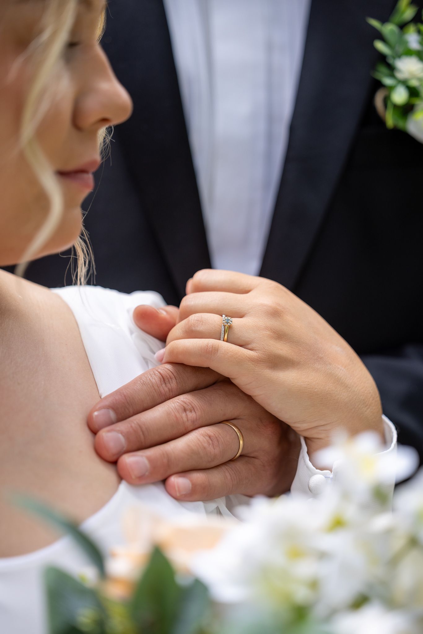 Bride and groom holding hands showing wedding rings, with white flowers in the foreground.