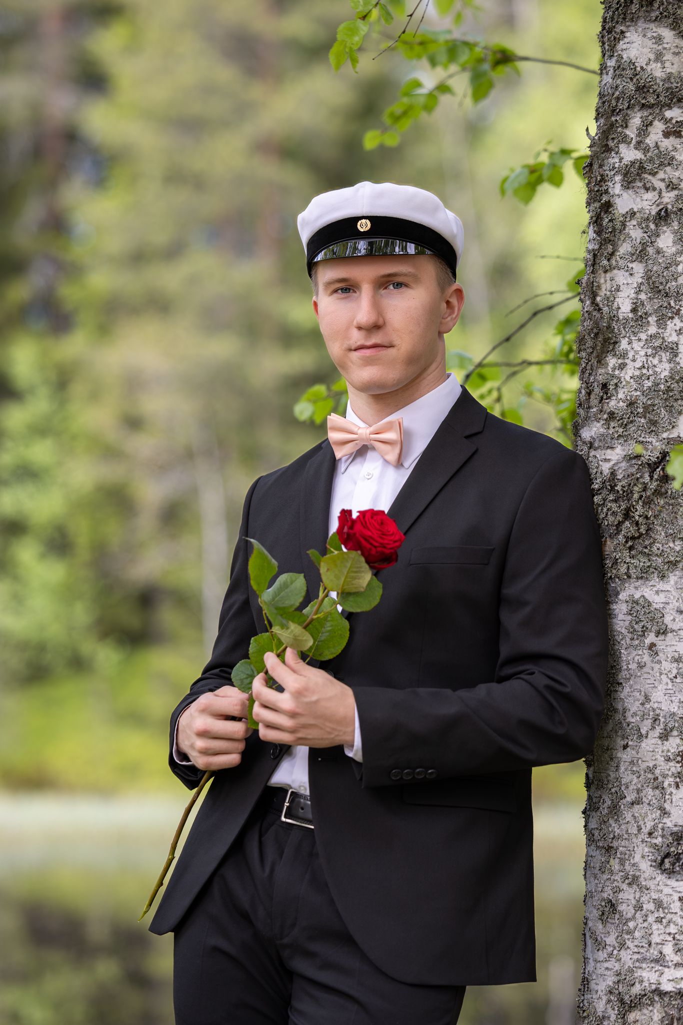 Young man in black suit, bow tie, and white cap holds a red rose while leaning against a birch tree outdoors.