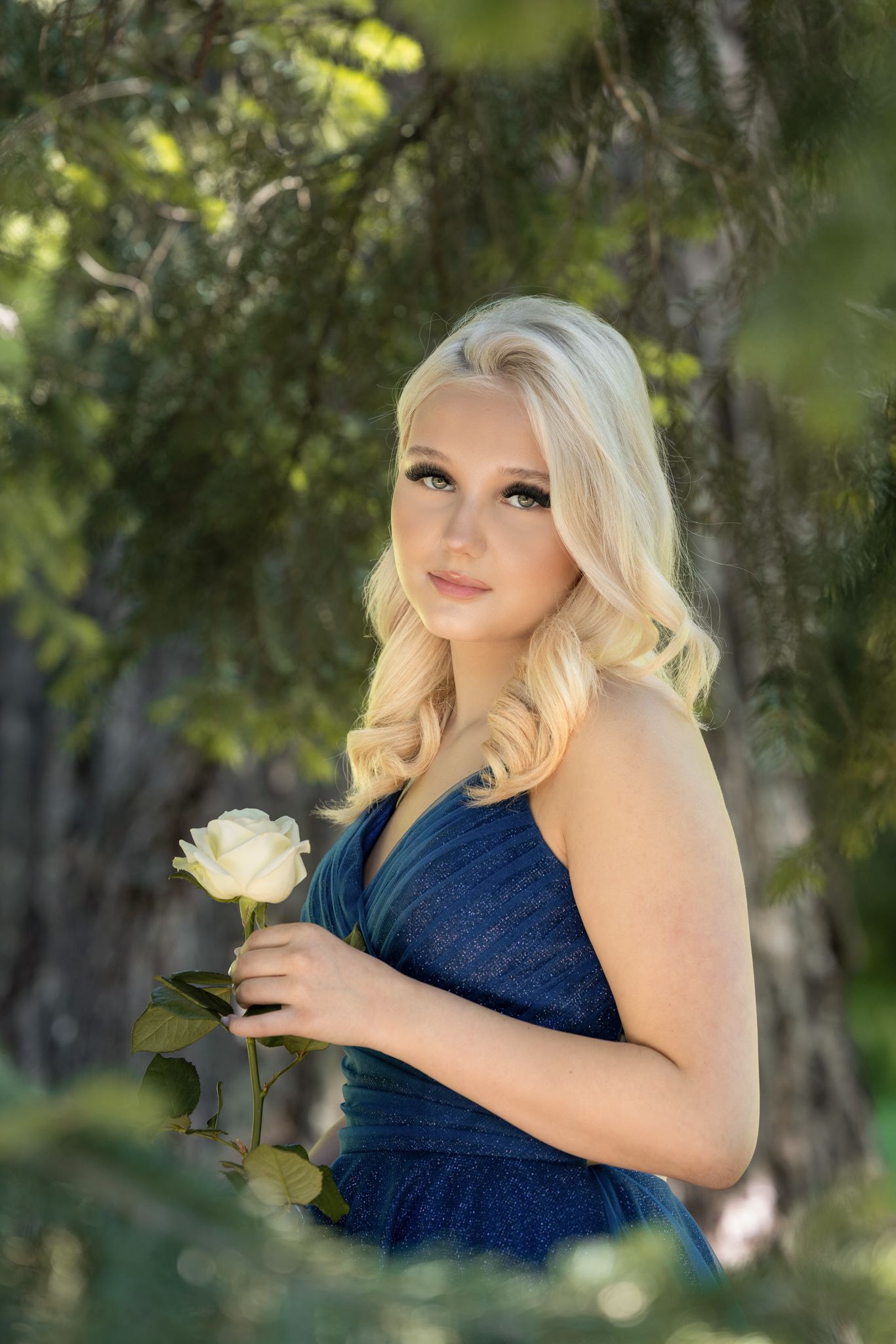 Young woman in navy blue dress holds white rose while posing outdoors in natural setting with soft lighting.