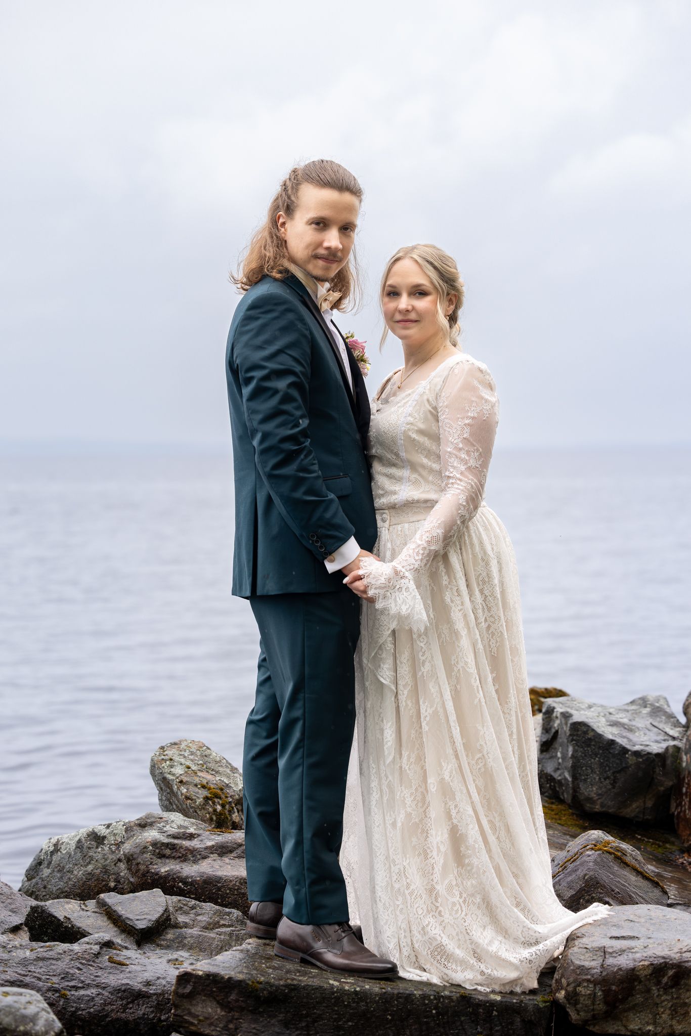 Bride and groom in wedding attire stand on rocky shoreline holding hands with overcast sky and ocean behind them.