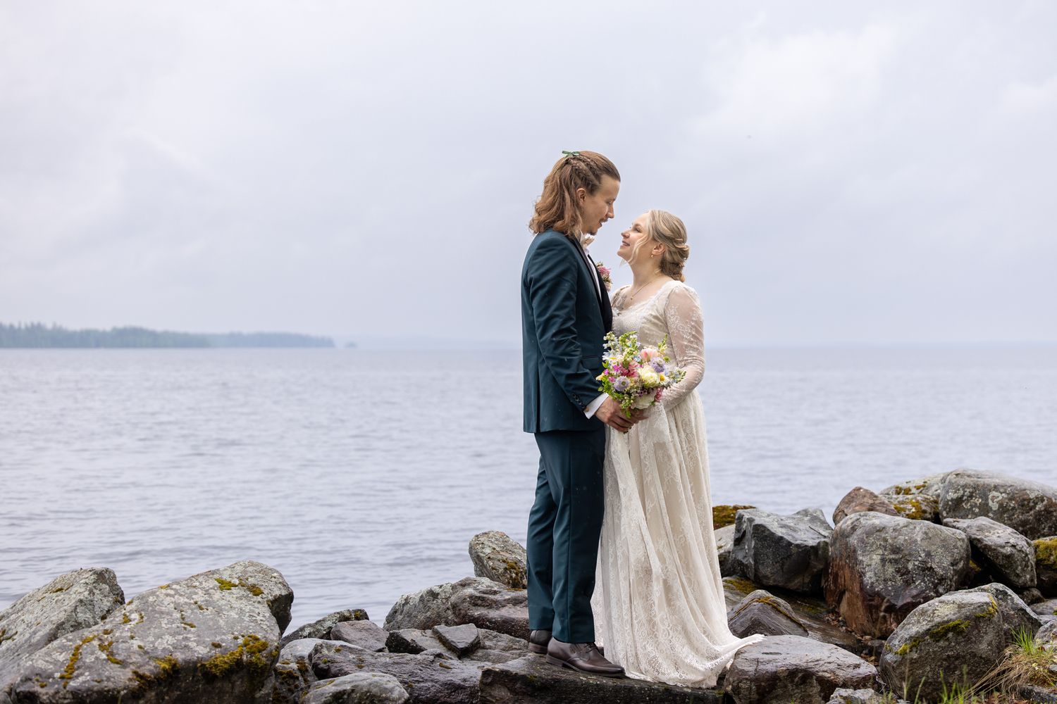 Bride and groom stand on rocky shoreline in wedding attire, sharing an intimate moment with misty ocean backdrop.