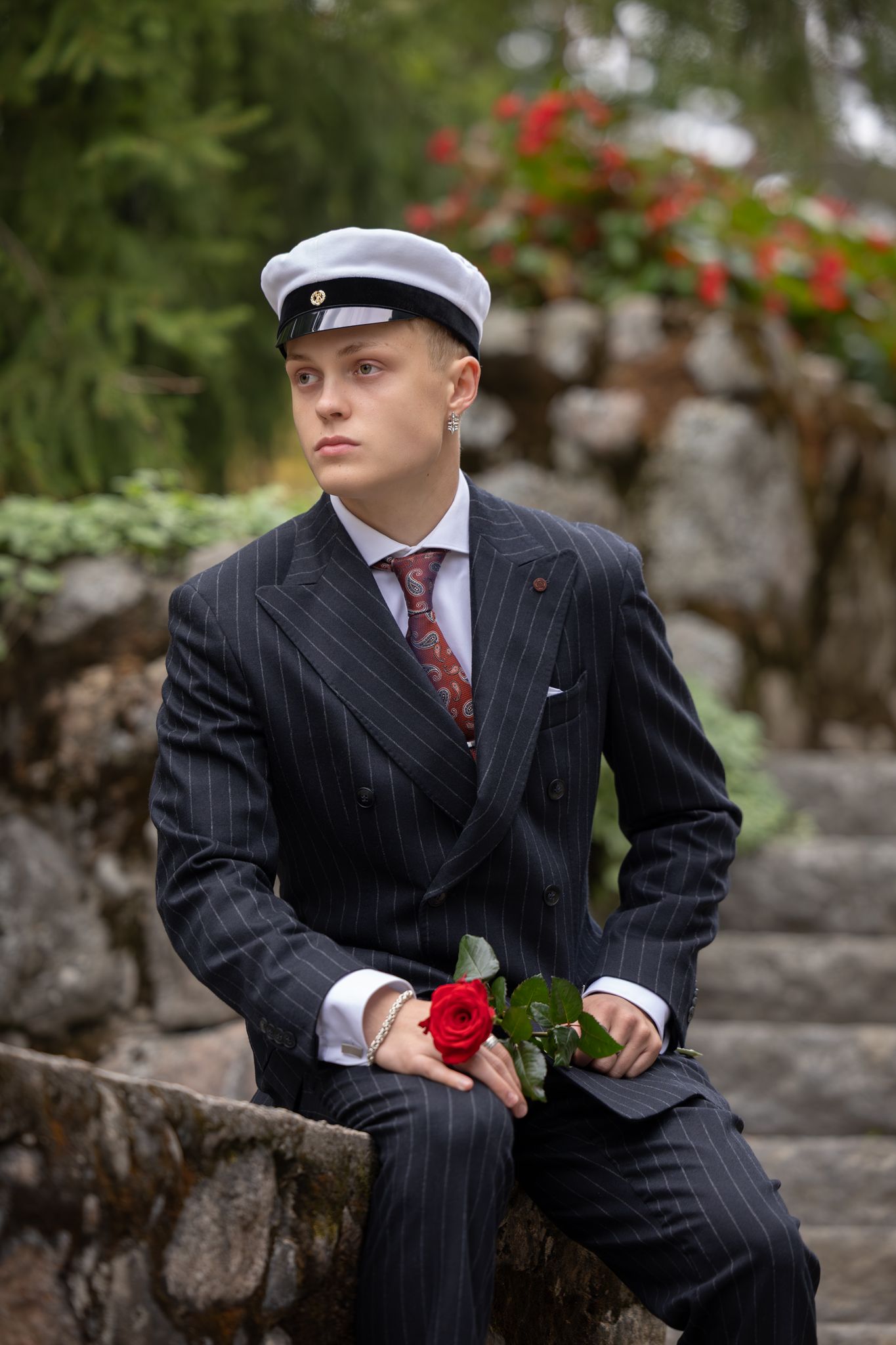 Young man in pinstripe suit and white cap holds a red rose while sitting on stone steps outdoors.