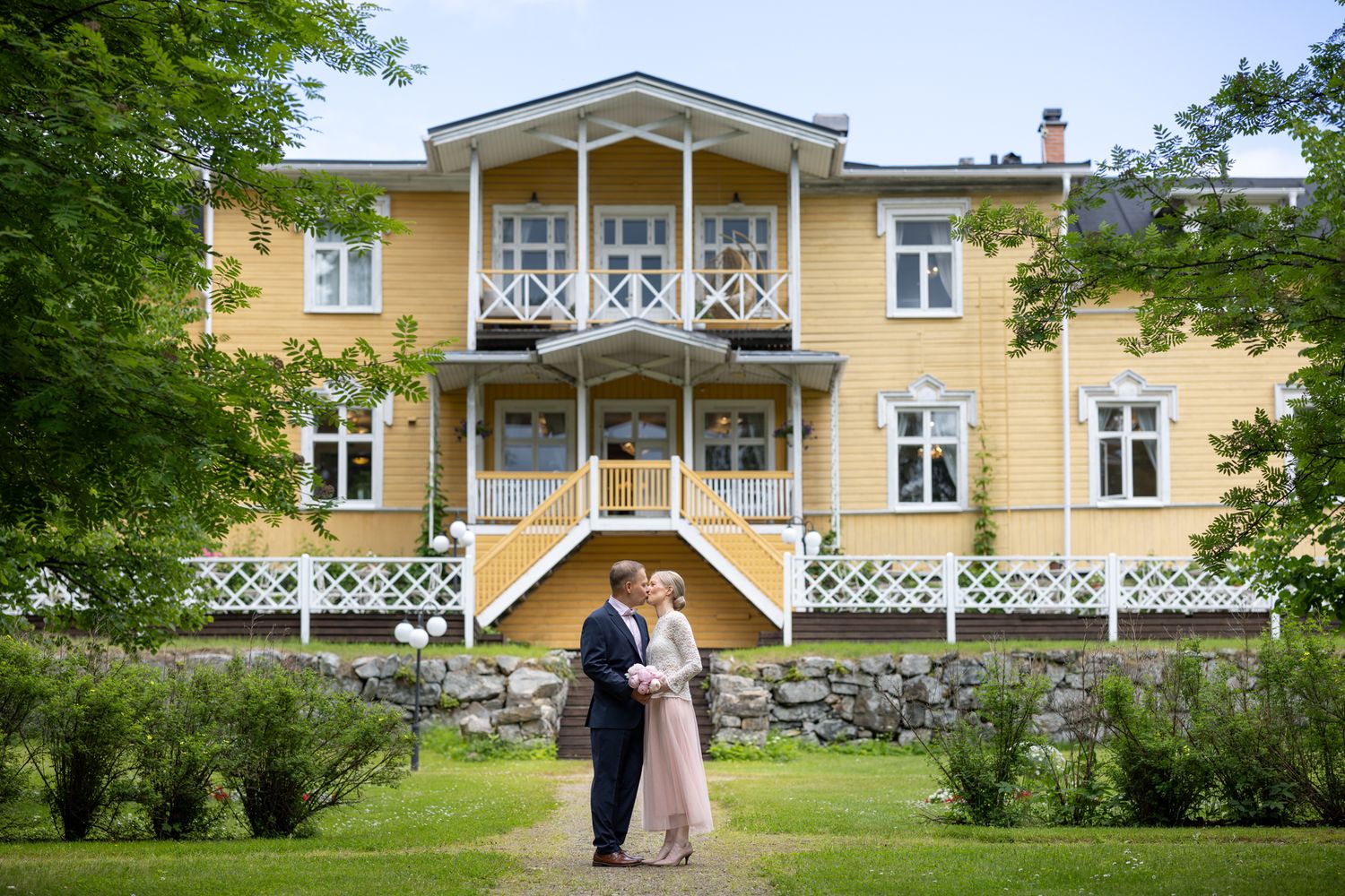 Couple embracing in front of a grand yellow wooden manor house with white balconies and lush green gardens.