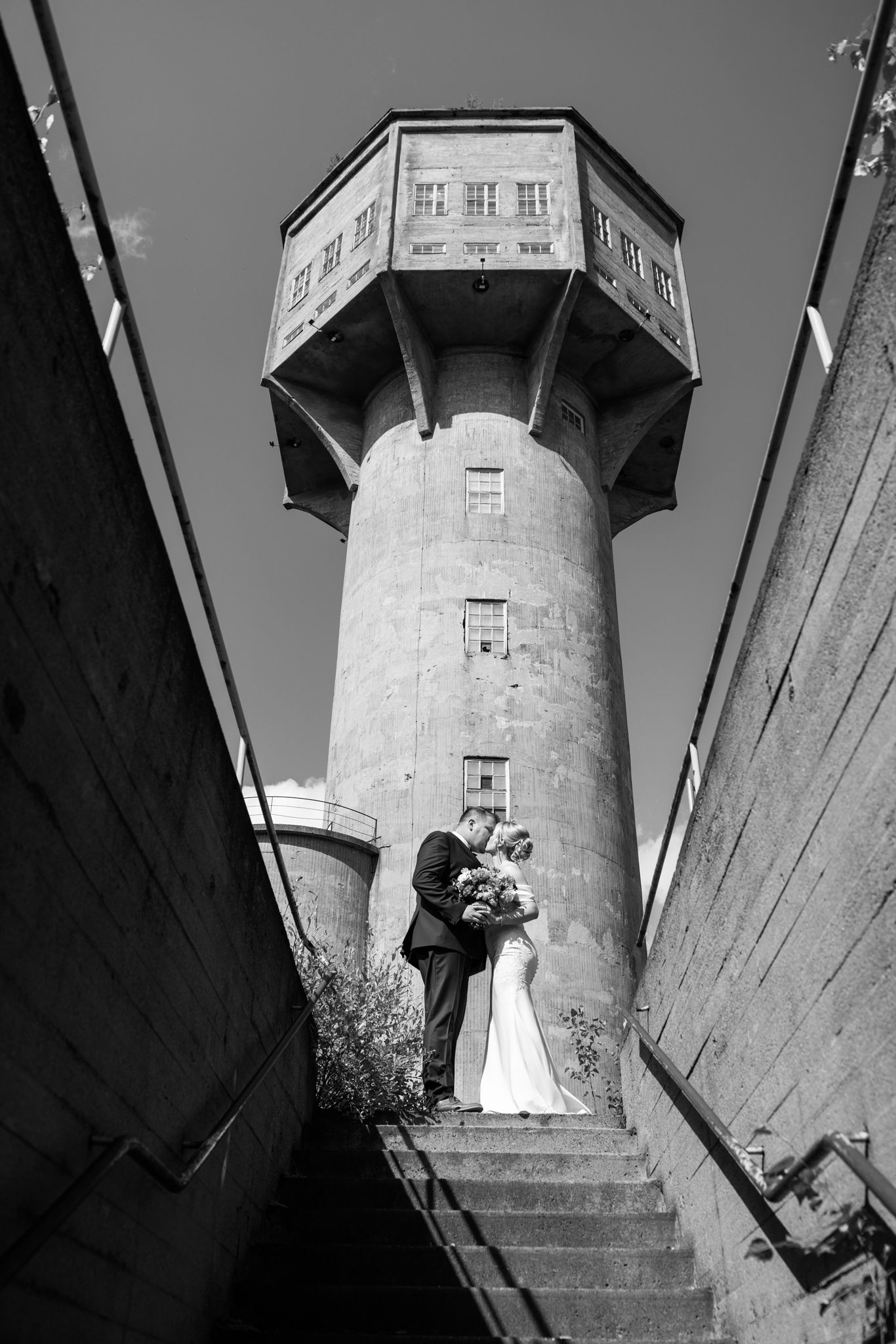 Black and white wedding photo of couple kissing on steps before a tall stone water tower.