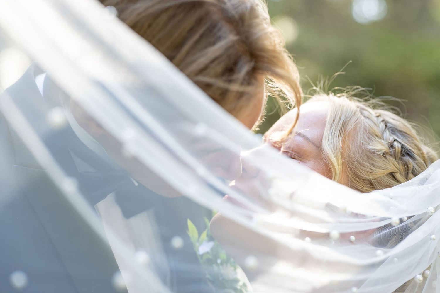 Two blonde women share a tender embrace under a flowing white bridal veil outdoors in soft natural light.