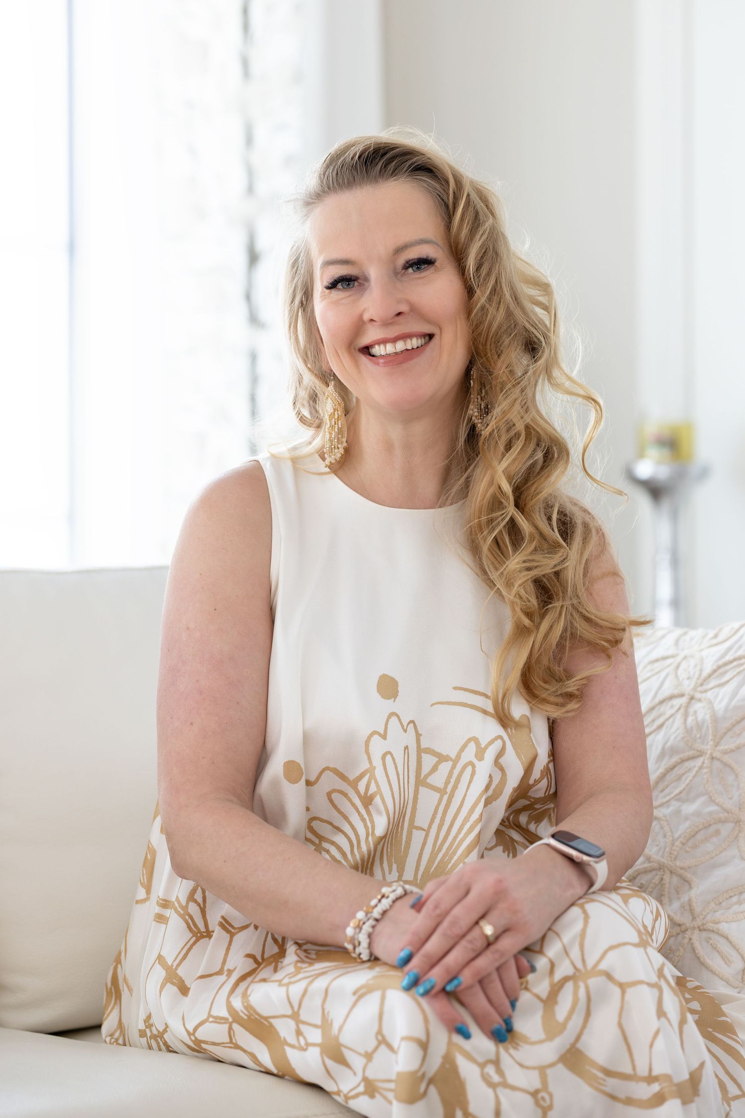 Smiling blonde woman in white and gold floral dress sitting on a sofa in a bright, elegant room.