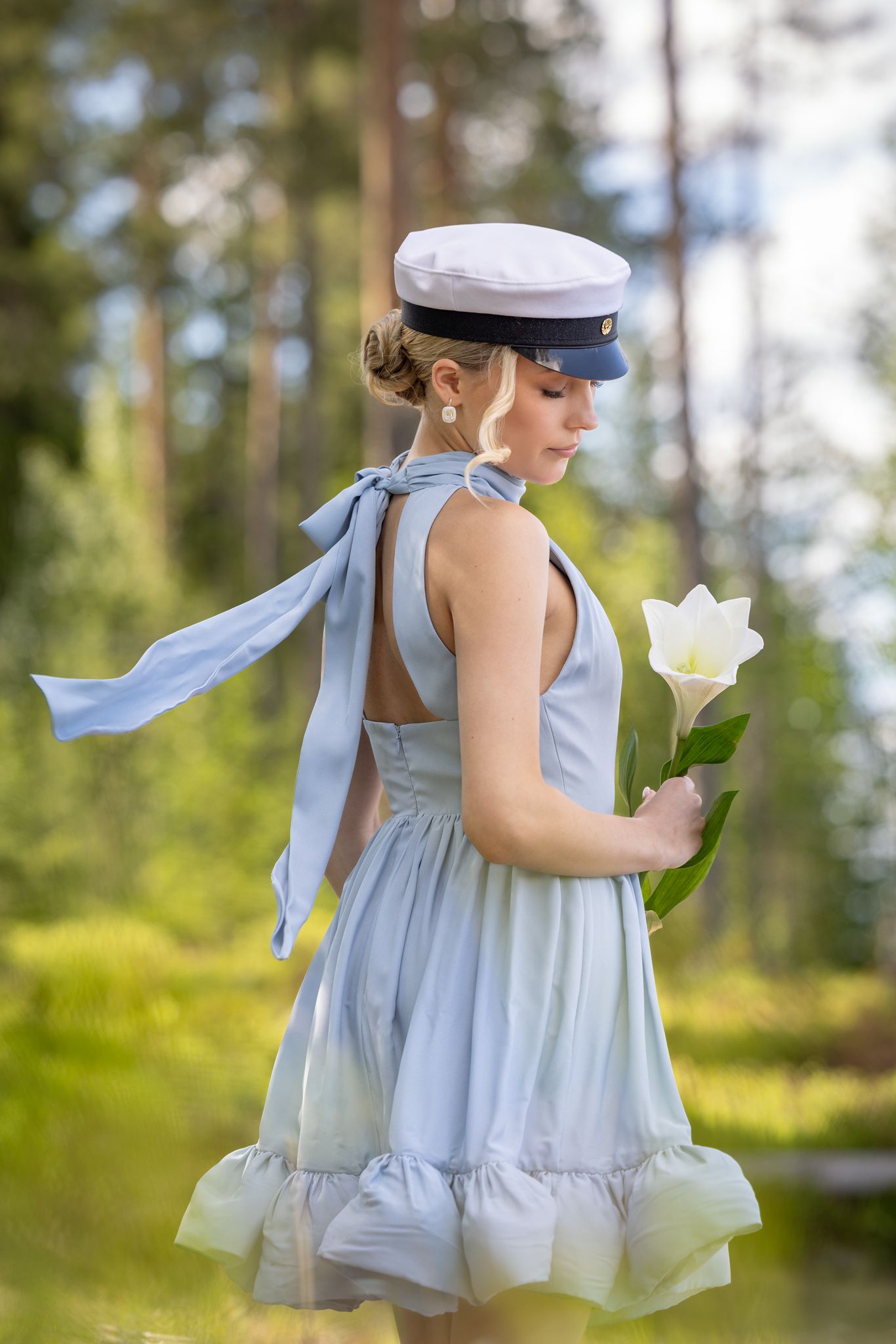 Young woman in light blue backless dress and white cap holds a white tulip in a sunlit forest setting.
