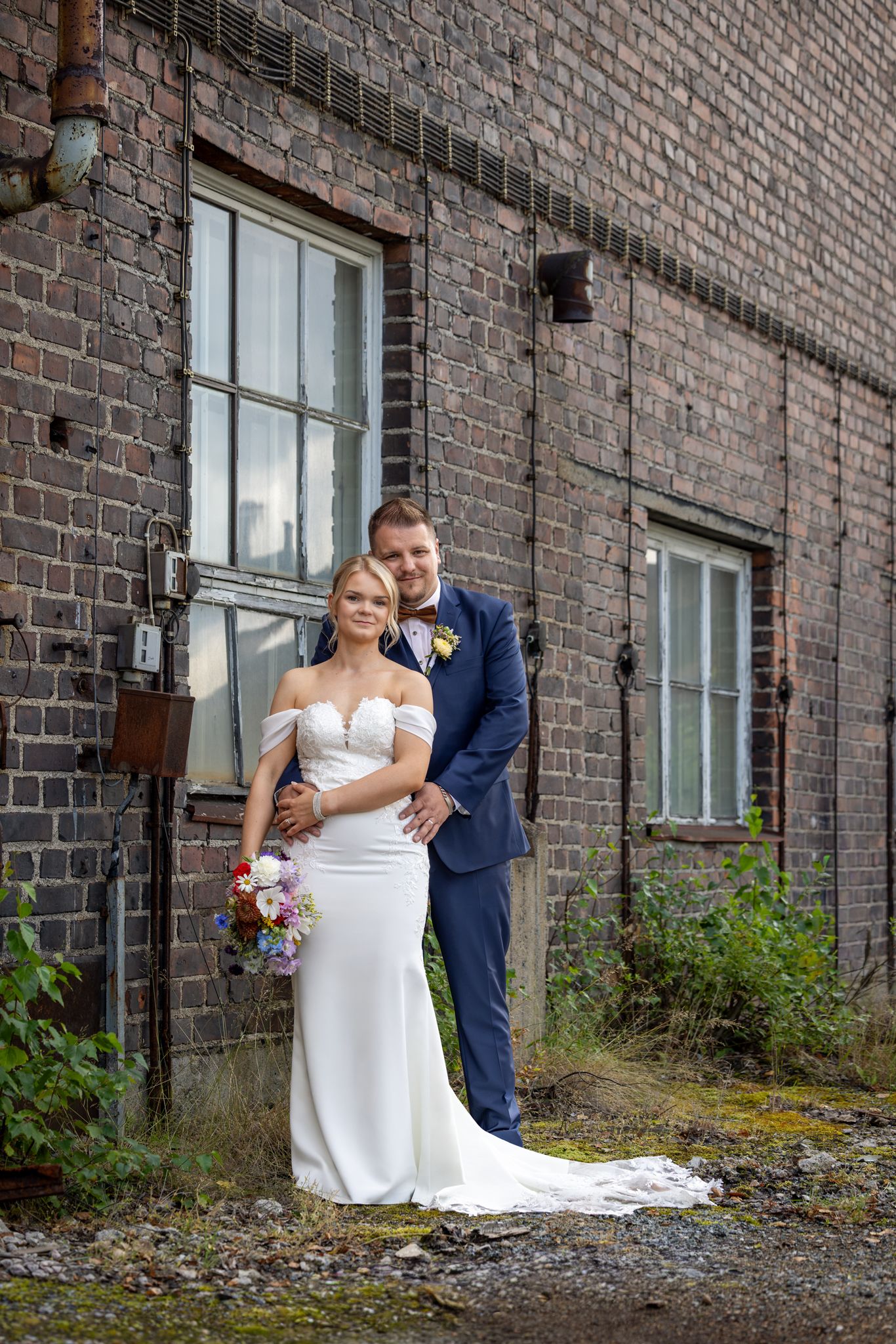 Bride in white gown and groom in navy suit pose together outside a rustic brick building on their wedding day.
