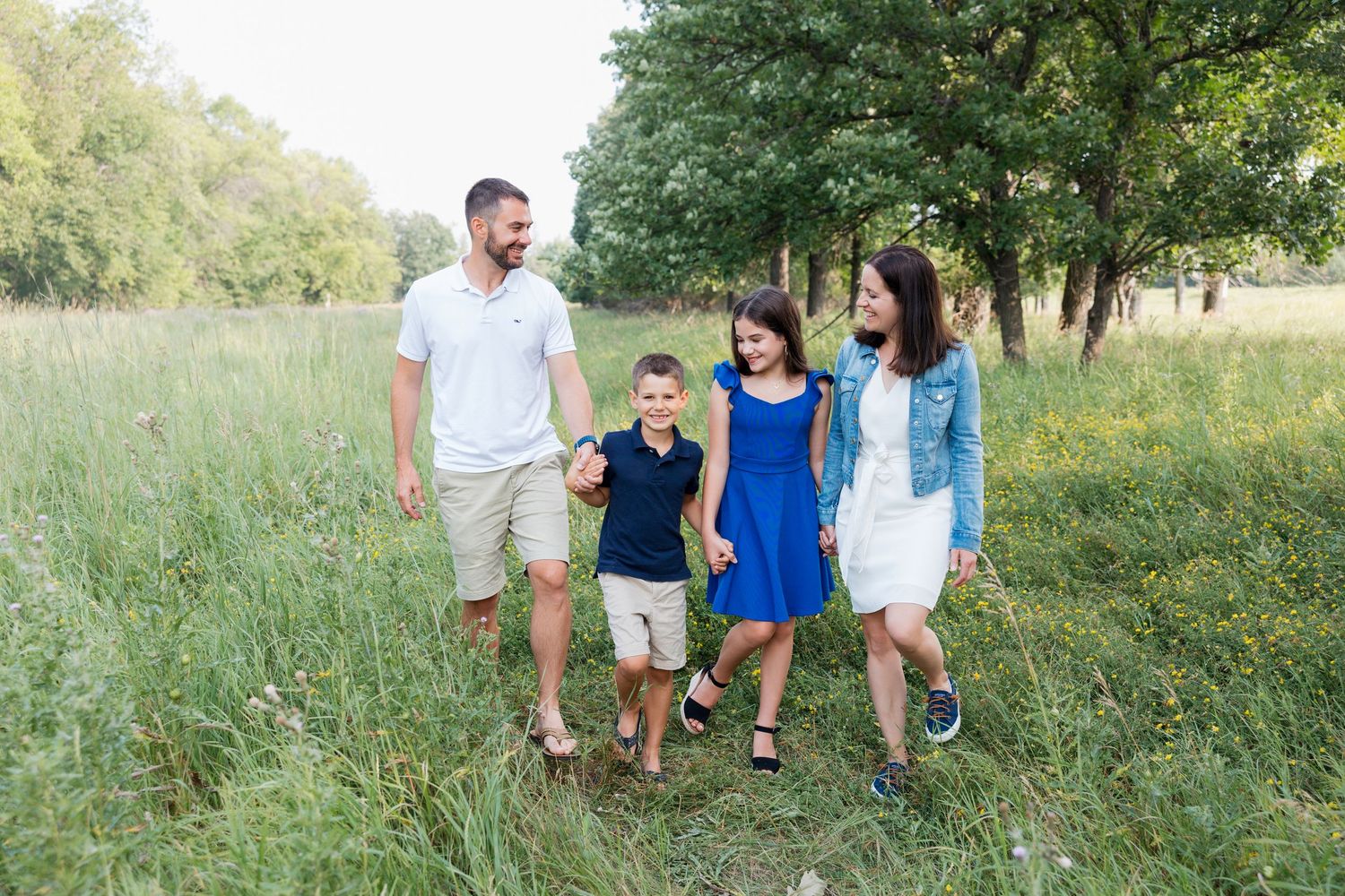 A family walks together through a sunny meadow with tall grass and trees in the background on a summer day.