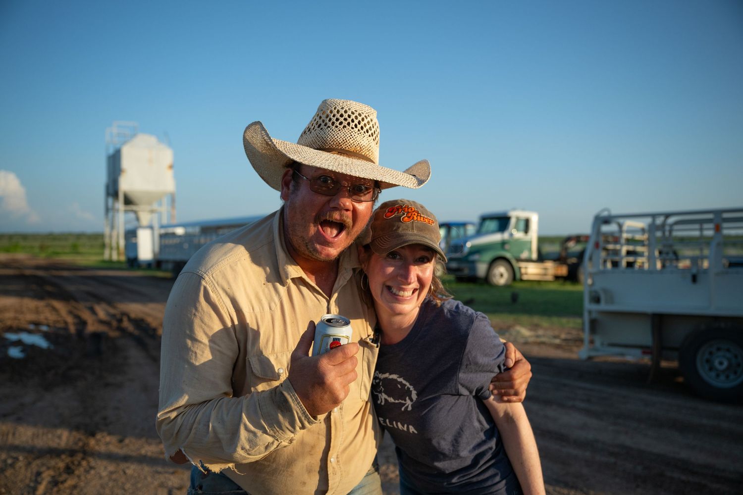 Two people wearing hats smile together outdoors with trailers and a white water tower visible in background.