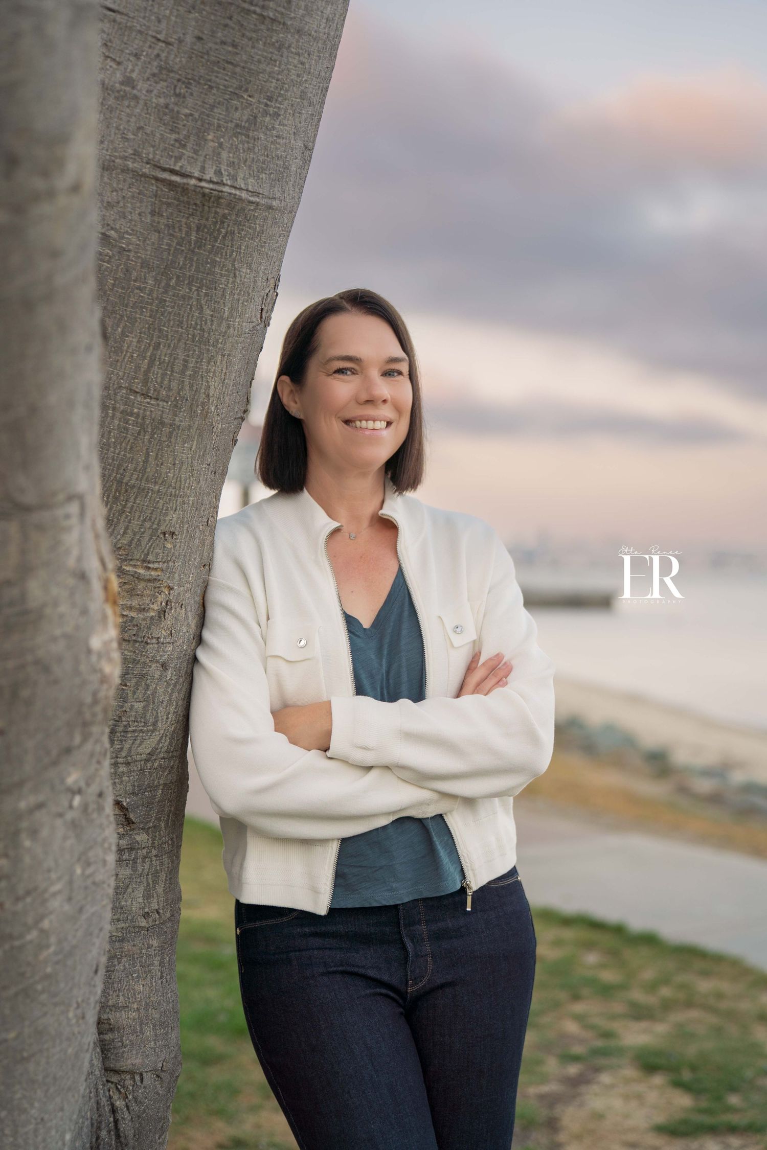 Professional outdoor portrait of someone in a white cardigan leaning against a tree at sunset.