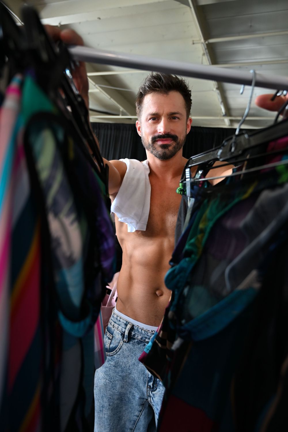 Athletic person in a white tank top and acid wash jeans poses between hanging garments in a wardrobe area.