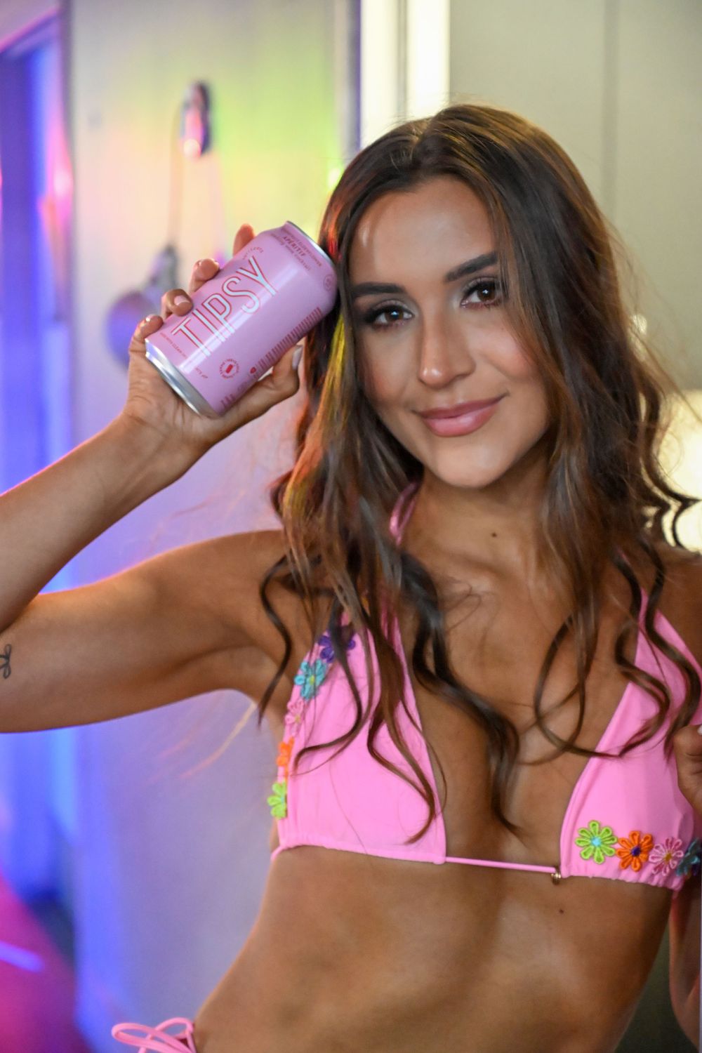 Athletic woman in pink floral bikini top holding pink drink can against colorful background.