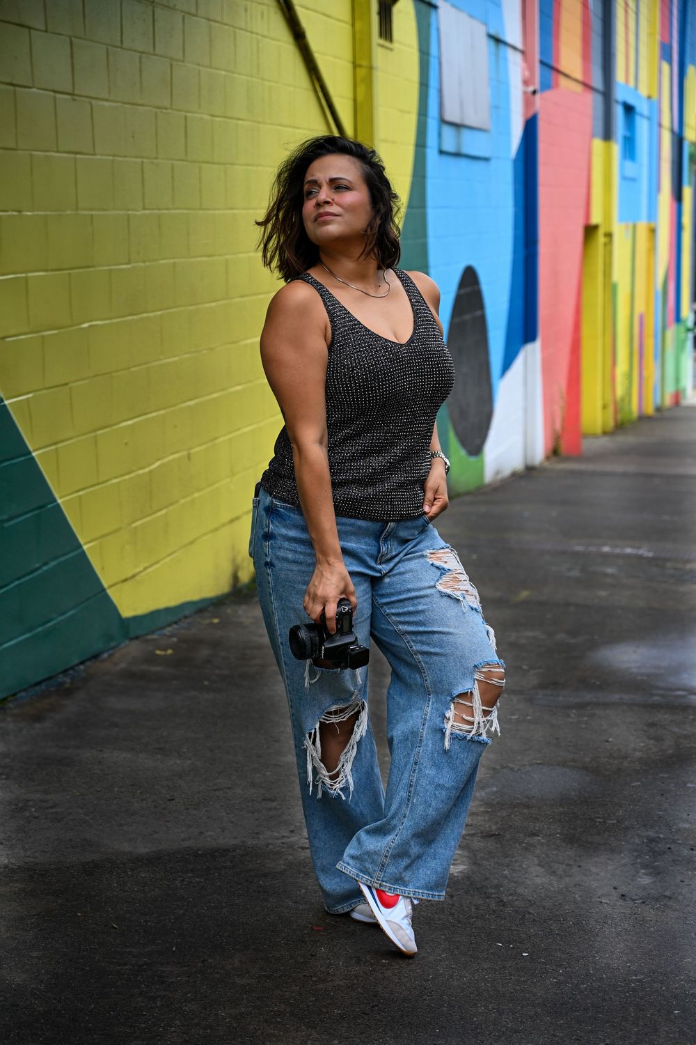 Person in distressed jeans and tank top poses confidently against colorful geometric mural wall on urban street.