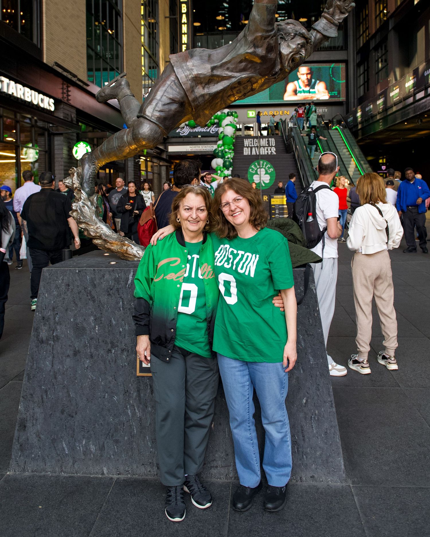Fans wearing green Boston Celtics jerseys pose by a statue outside TD Garden before a basketball game in Boston.