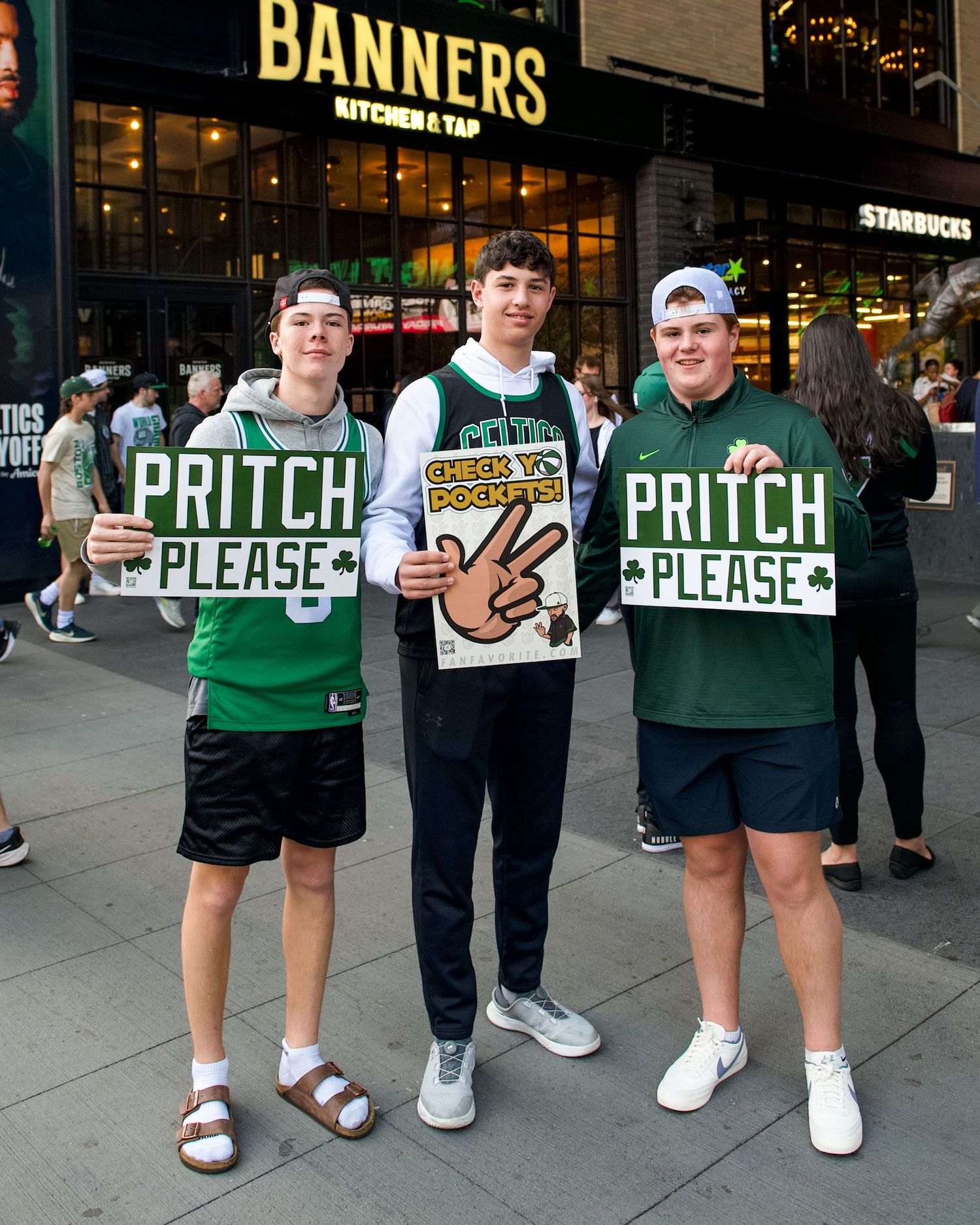 Three basketball fans hold signs that read 'PRITCH PLEASE' outside Banners restaurant in green jerseys.
