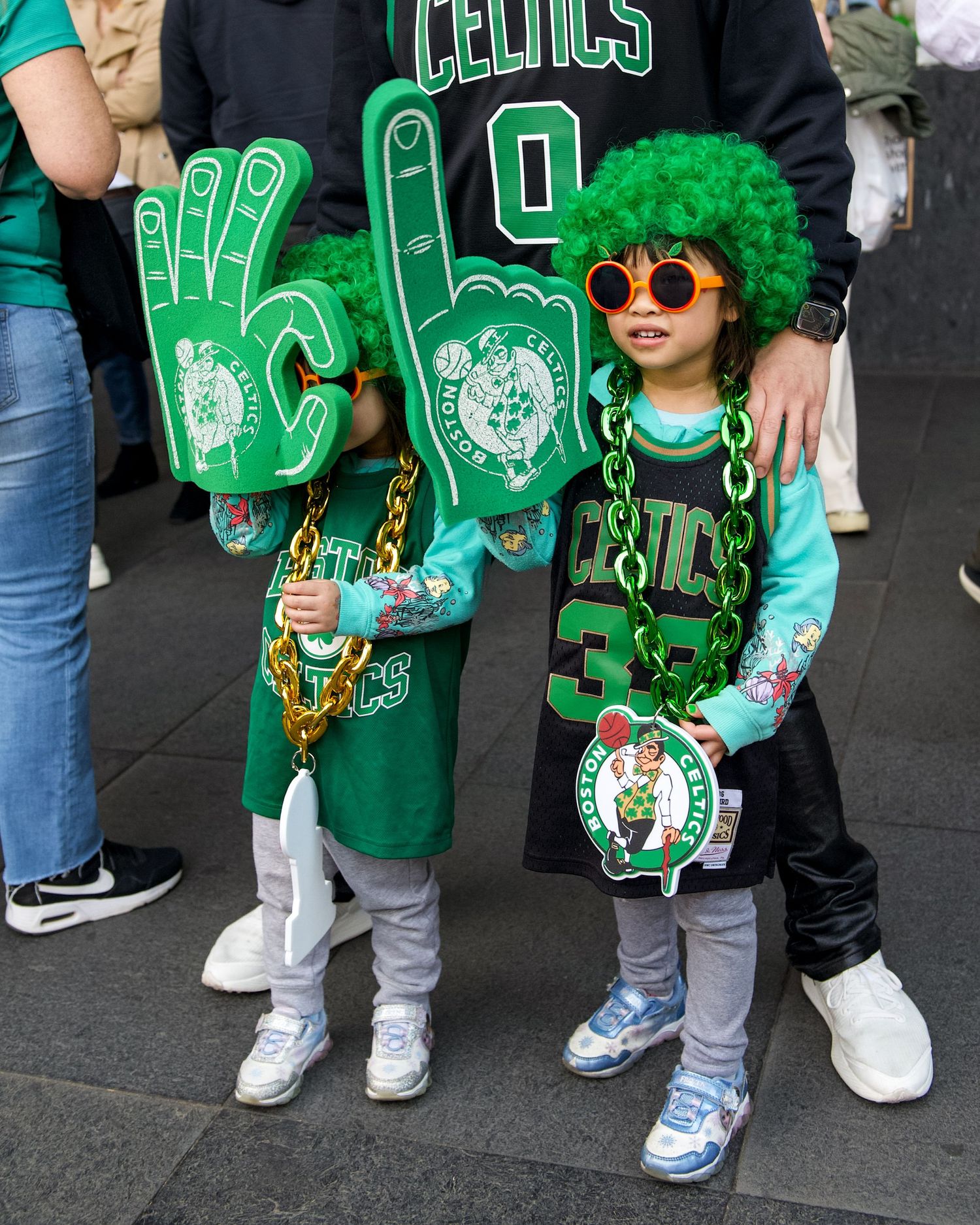 Two young Boston Celtics fans wear matching green team gear with foam fingers and St. Patrick's Day accessories.