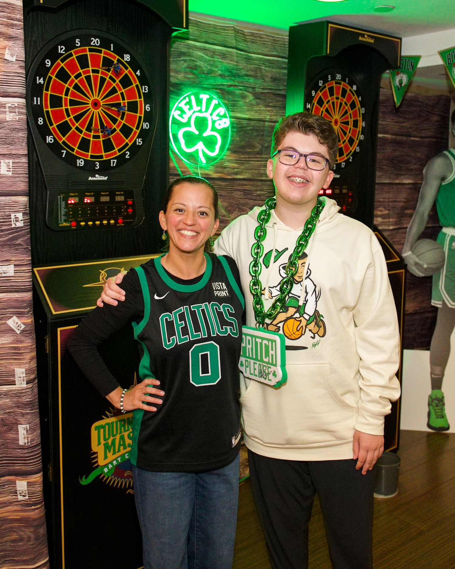 Two people pose together in front of electronic dartboards with Celtic-themed decor and green lighting in a game room setting.