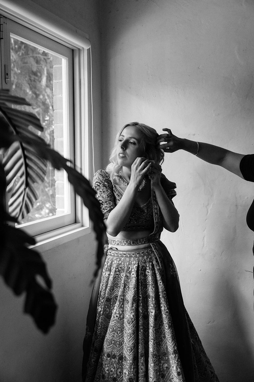 Black and white photo of someone in a vintage dress being styled near a window with dramatic natural light.