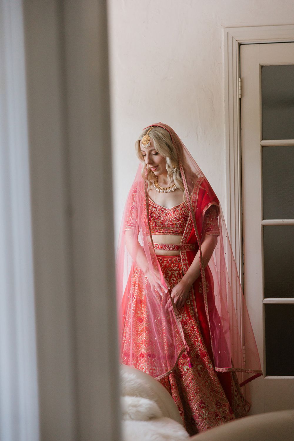 Traditional Indian bride wearing an elegant red lehenga with intricate embroidery and a sheer veil stands by a doorway.