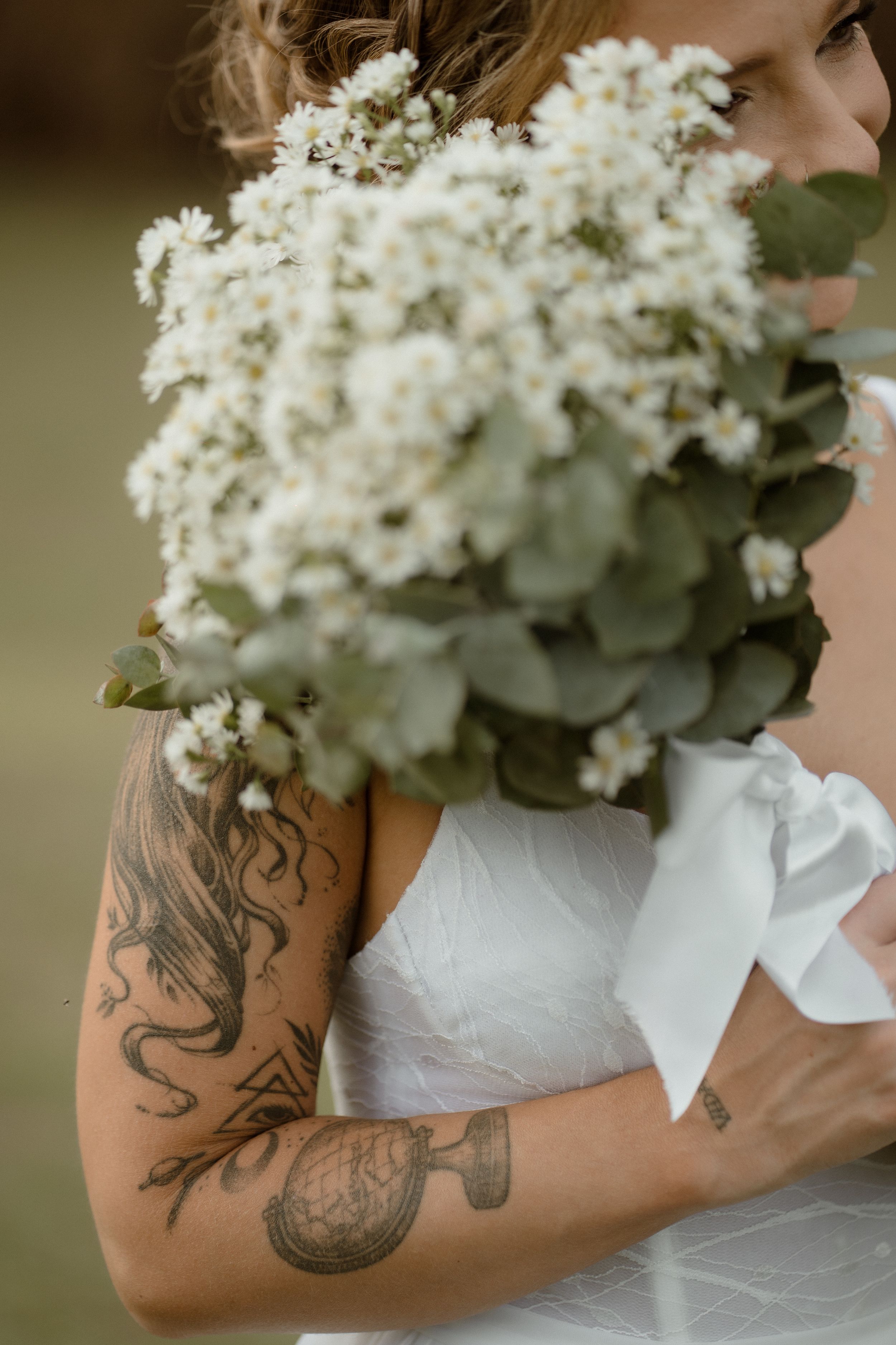 White bridal bouquet held against white dress with artistic tattoo visible on arm.