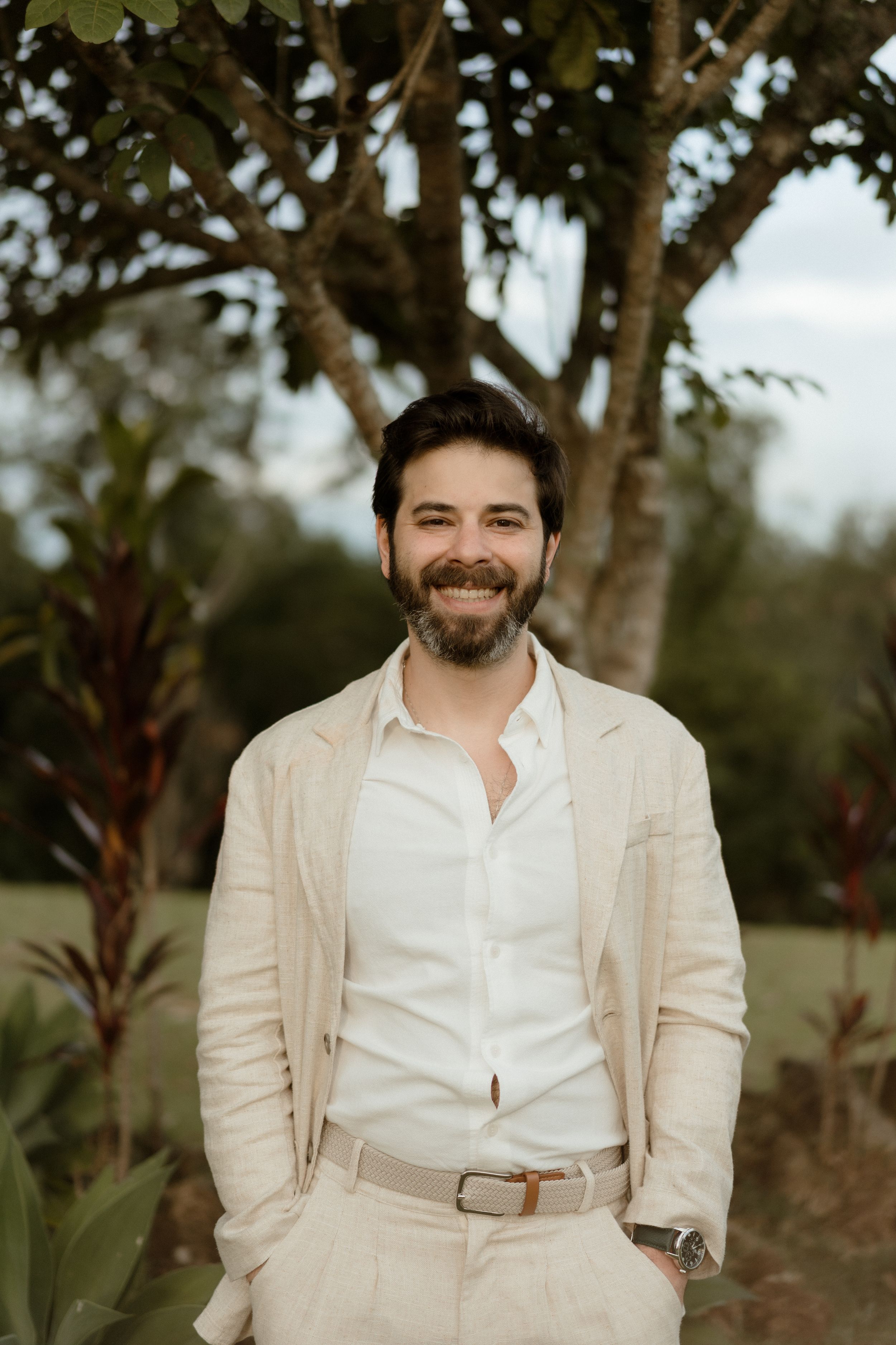Portrait of person in cream suit and white shirt standing against garden background.