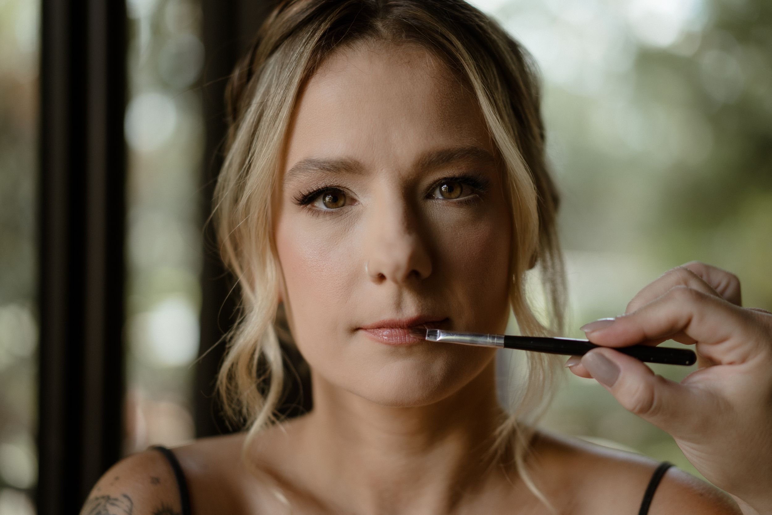 Makeup artist applies cosmetics during a wedding preparation with natural window lighting in the background.