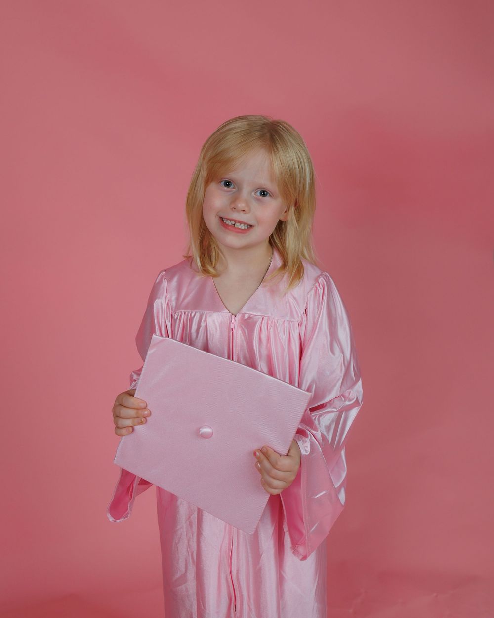 Child wearing pink graduation gown holds a blank paper against pink background.