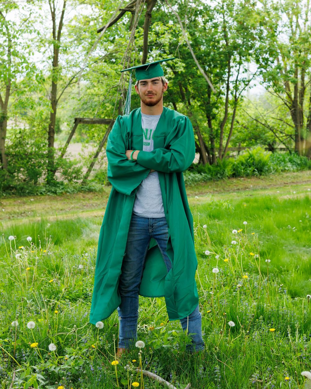 Graduate in green cap and gown stands confidently in a sunny meadow with wildflowers and trees in the background.