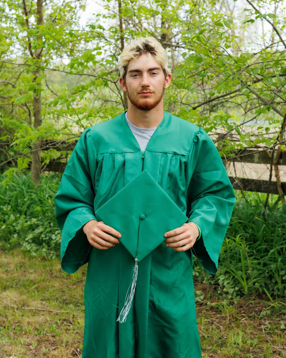 A person wearing a bright green graduation gown with tassel stands outdoors against a leafy green natural background.