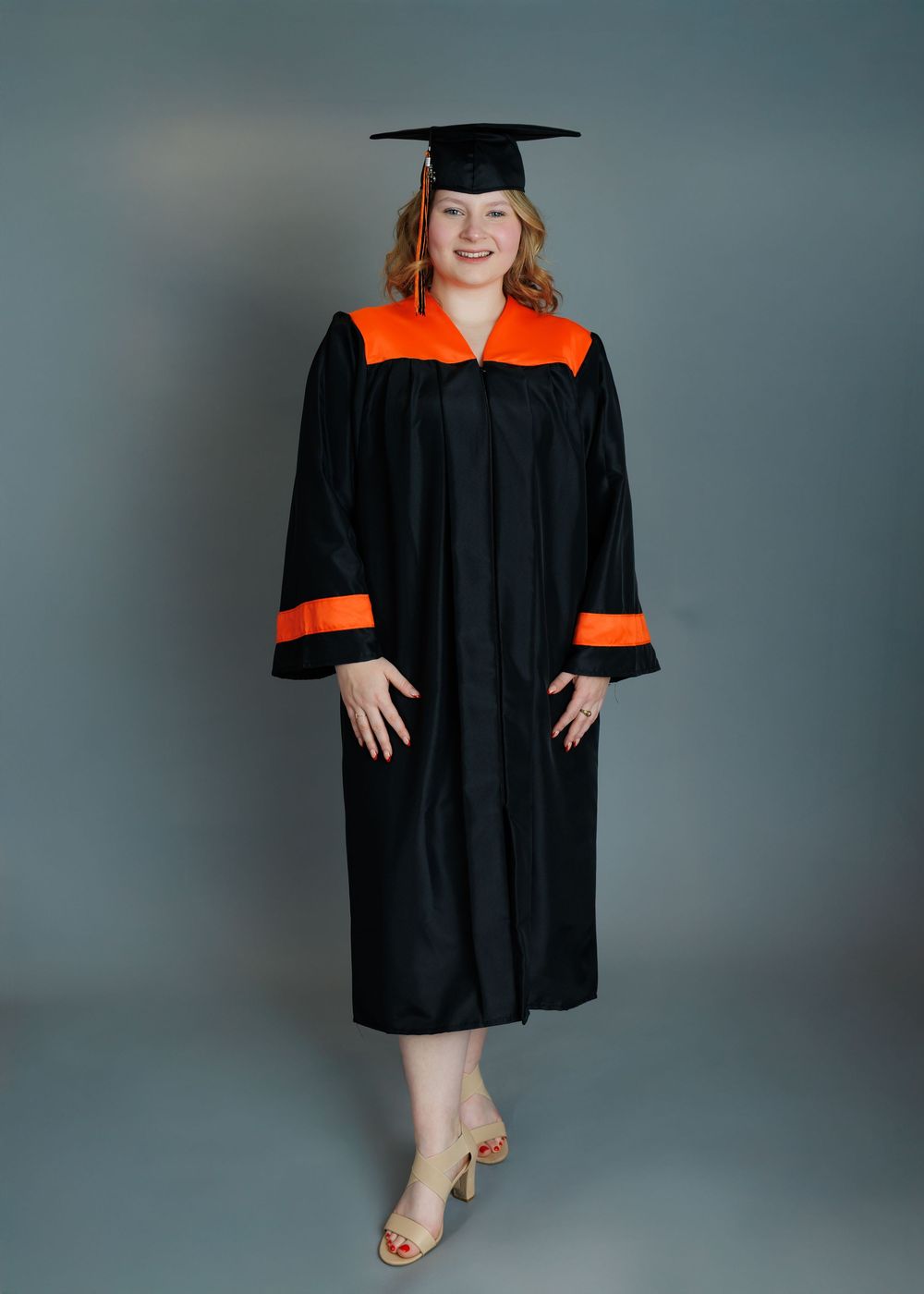 Graduate in black cap and gown with orange accents poses confidently in front of gray backdrop for graduation photos.