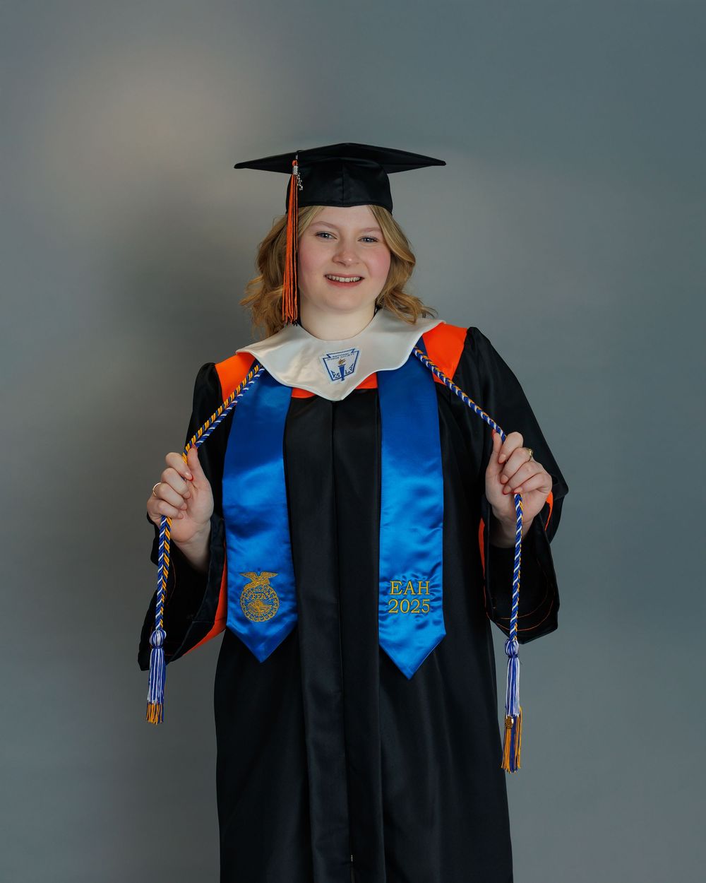 Graduate in black cap and gown with bright blue and orange stoles holds graduation cords during commencement ceremony.